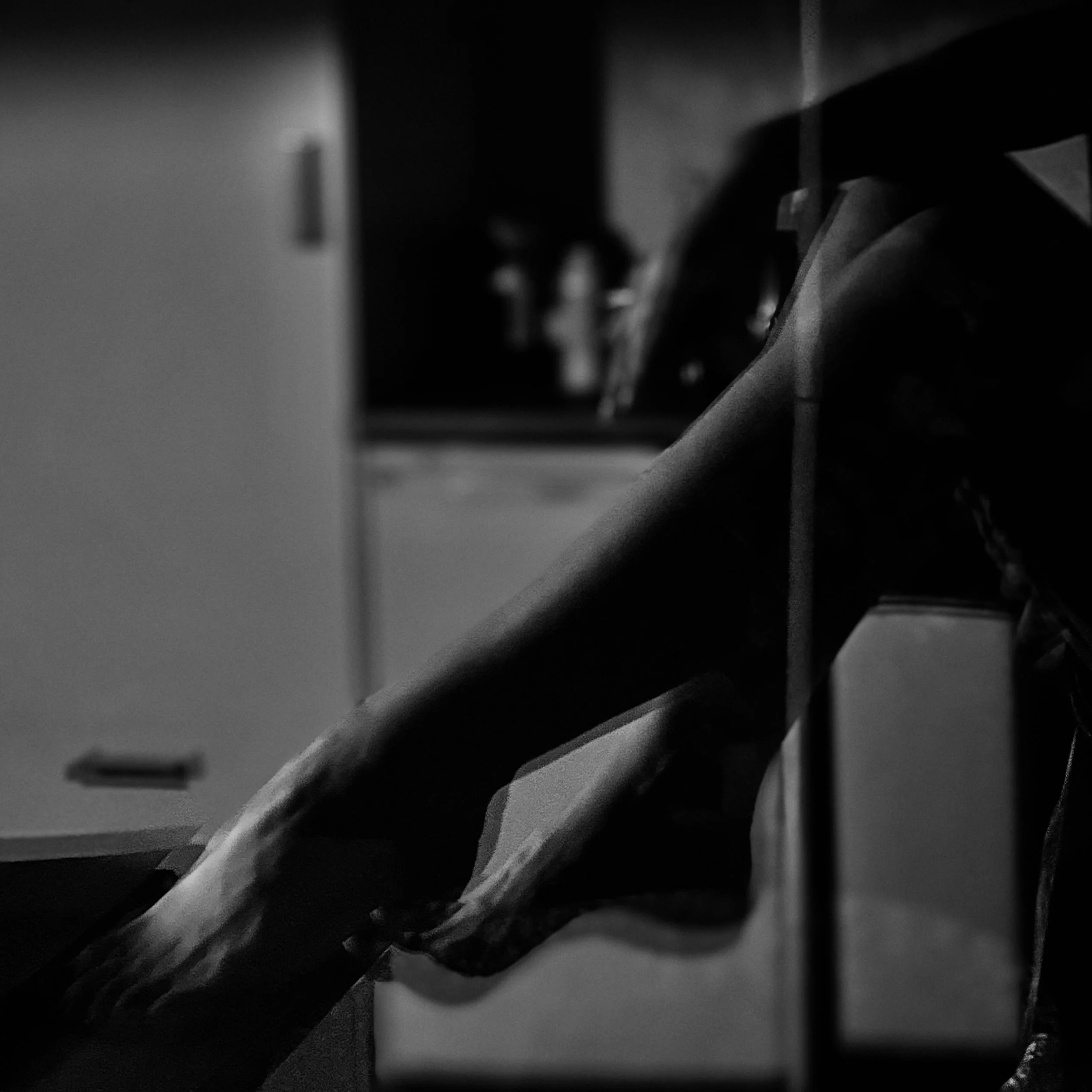 Black-and-white image of a person sitting indoors at night, legs crossed, partially lit, in a quiet kitchen setting.