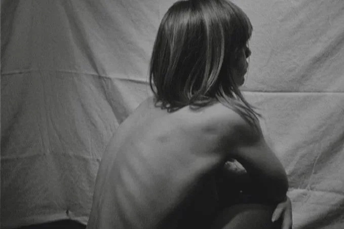 Black and white self-portrait of a woman seated with her back turned to the camera, exploring body posture, vulnerability, and sculptural form.