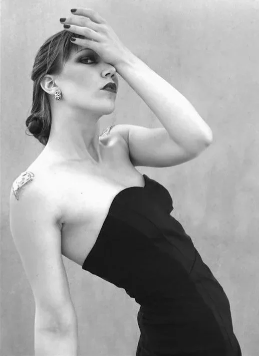 Black and white self-portrait of a woman wearing a strapless black dress, leaning back with one hand resting on her forehead.