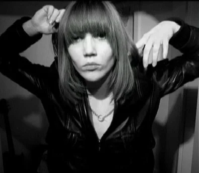 Color self-portrait of a woman with straight bangs covering her eyes, wearing a leather jacket, photographed indoors.