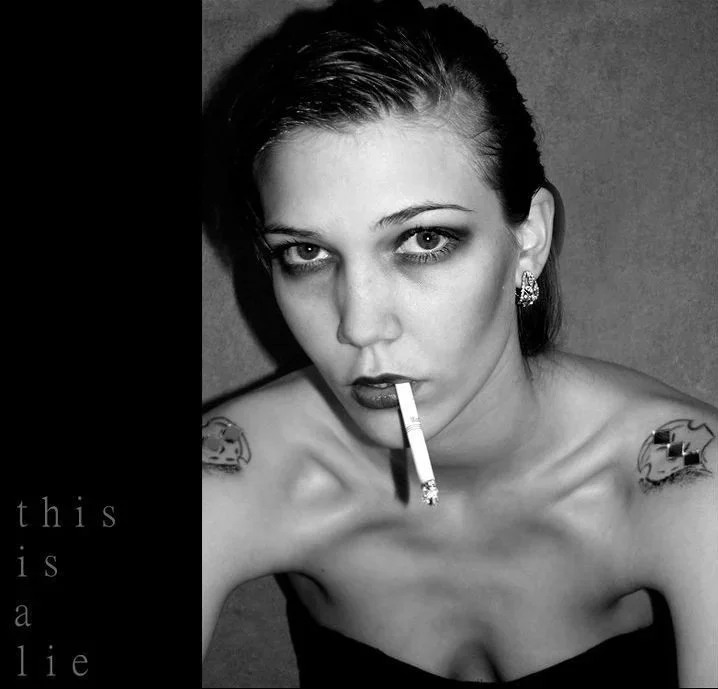 Black and white self-portrait of a woman with slicked-back hair wearing a strapless dress, smoking a cigarette and looking directly at the camera.