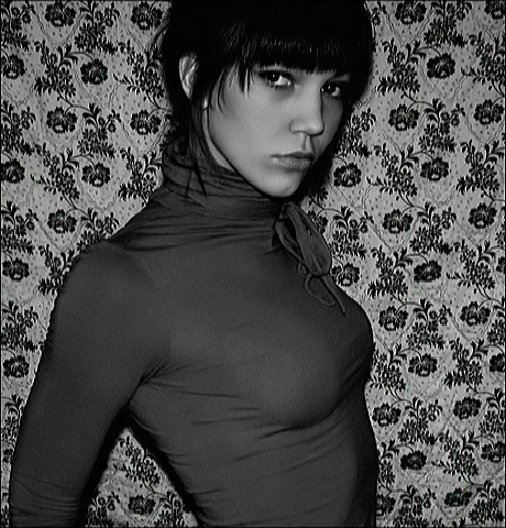 Black-and-white self-portrait of a woman against patterned wallpaper, direct gaze, intimate framing