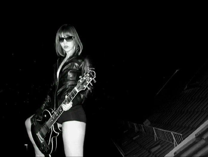 Black and white self-portrait photograph of a woman standing on a rooftop at night, wearing a leather jacket and holding an electric guitar.