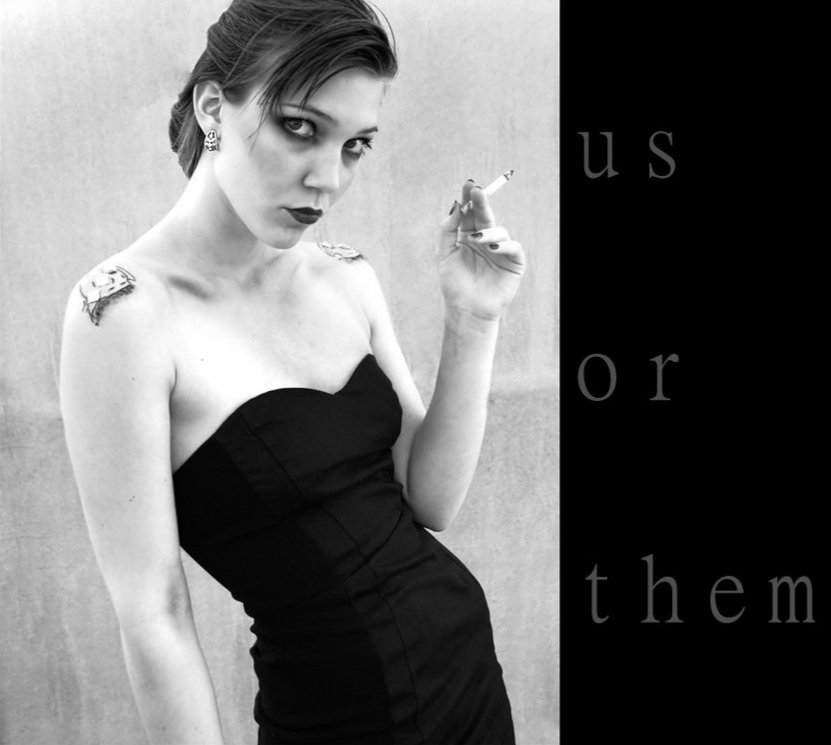 Black and white self-portrait of a woman wearing a strapless black dress, holding a cigarette and standing against a plain wall.
