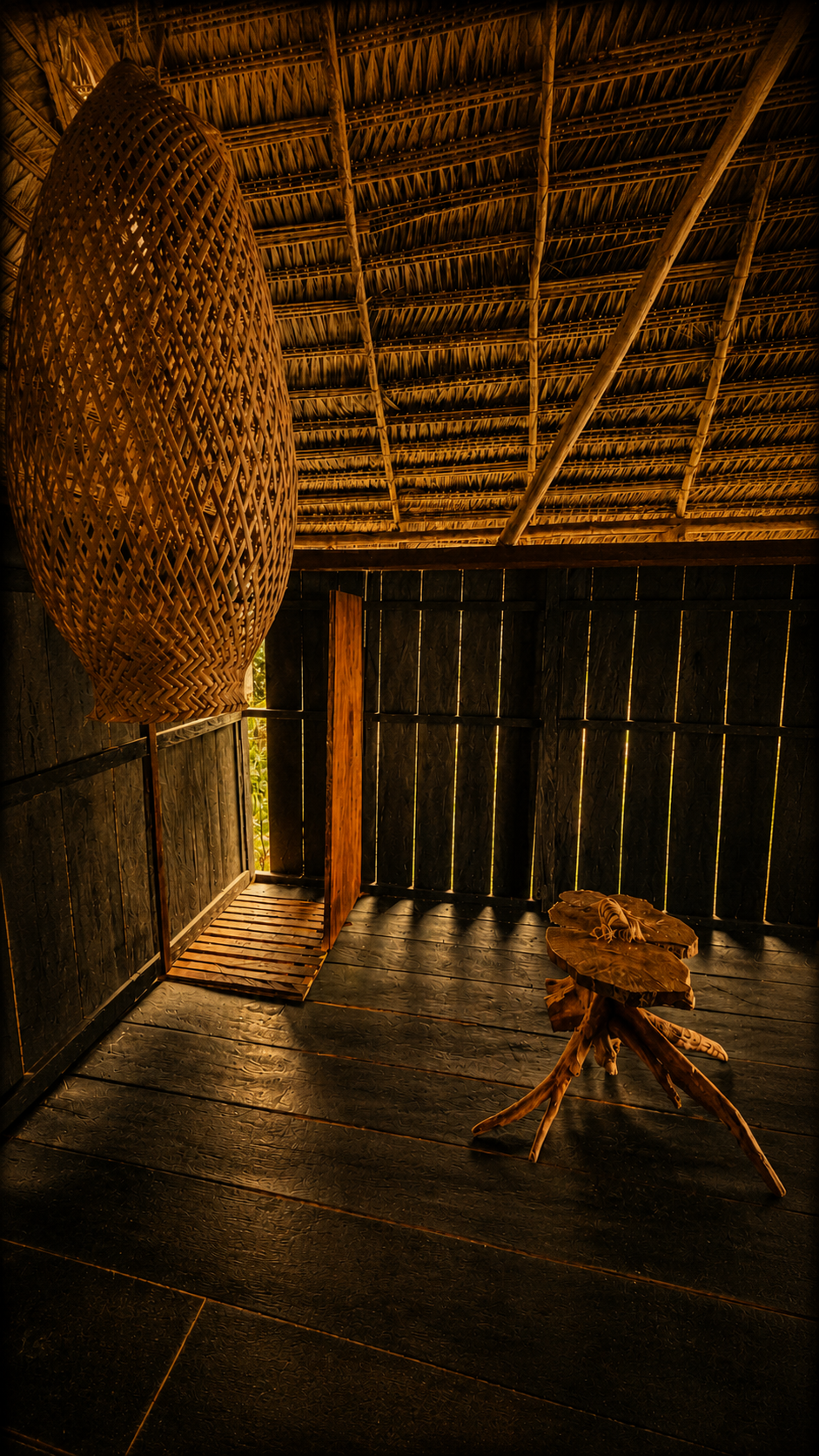 Interior de una cabaña rústica con paredes y piso de madera oscura, techo de paja y decoraciones de madera tallada. Un candelabro de mimbre colgado del techo.