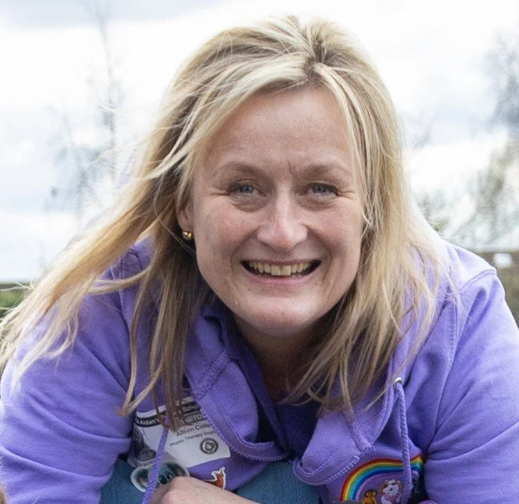 A woman with blonde hair smiling outdoors, wearing a purple jacket with patches and a rainbow badge.