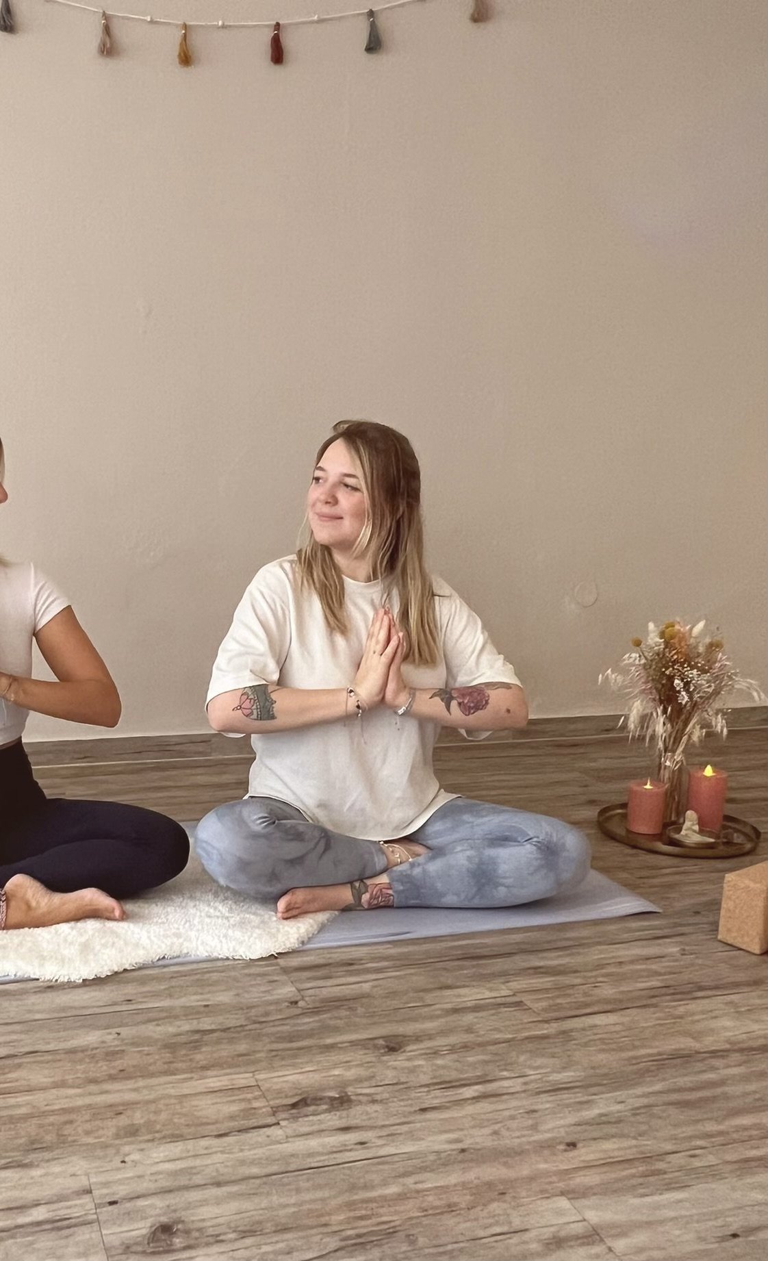 Eine junge Frau sitzt mit gekreuzten Beinen auf dem Boden, ihre Hände in Gebetshaltung, in einem Raum mit Kerzen und Dekoration, während sie Yoga oder Meditation macht.