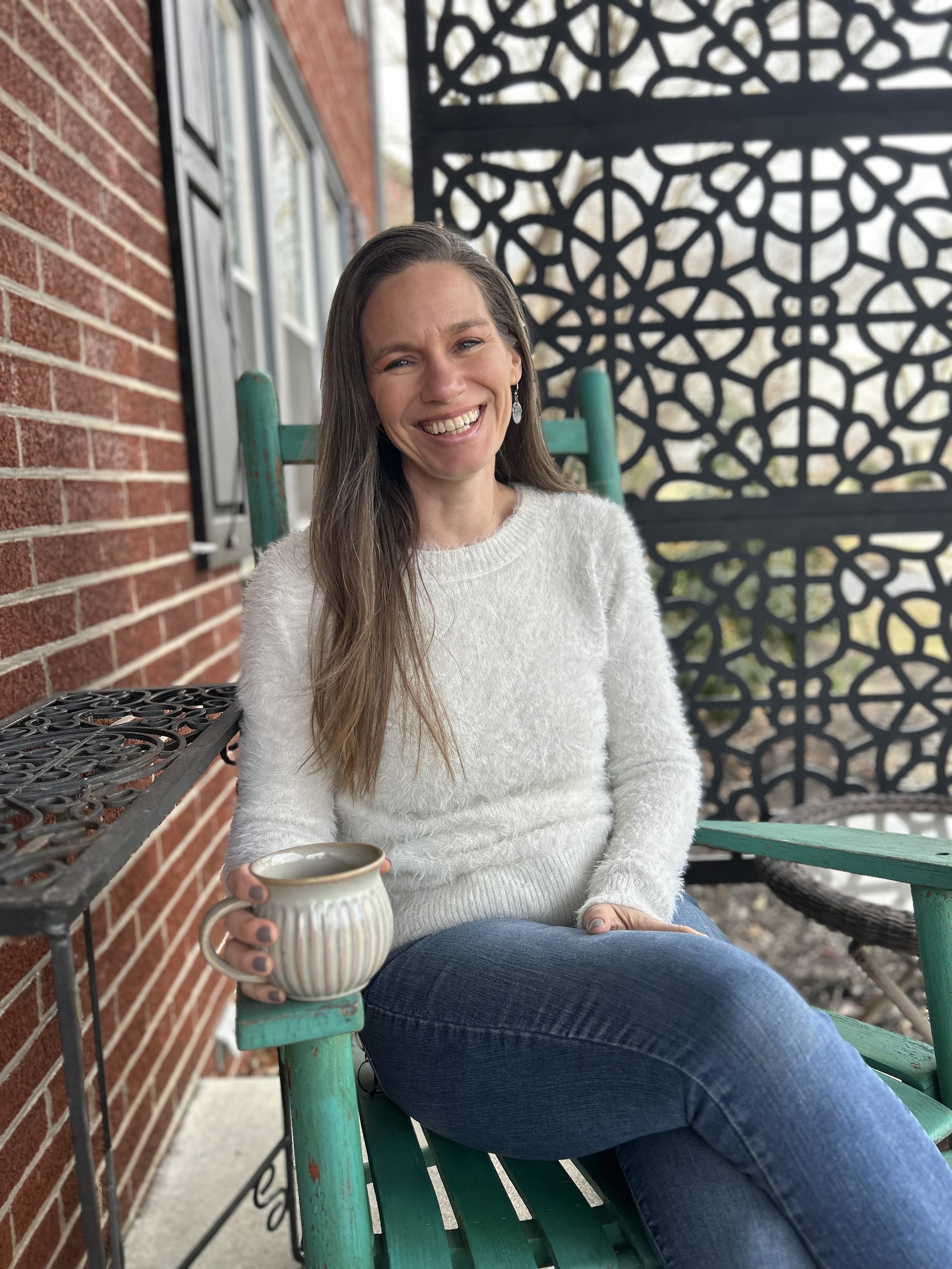 Me, a woman with long brown hair, wearing a white sweater and blue jeans, sitting on a teal bench on a porch, smiling and holding a mug. Behind me is a brick wall and a decorative black metal privacy screen.
