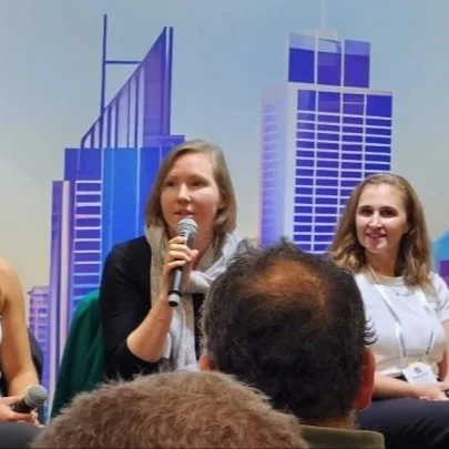 Two women speaking at a panel discussion with a cityscape backdrop, audience members listening.