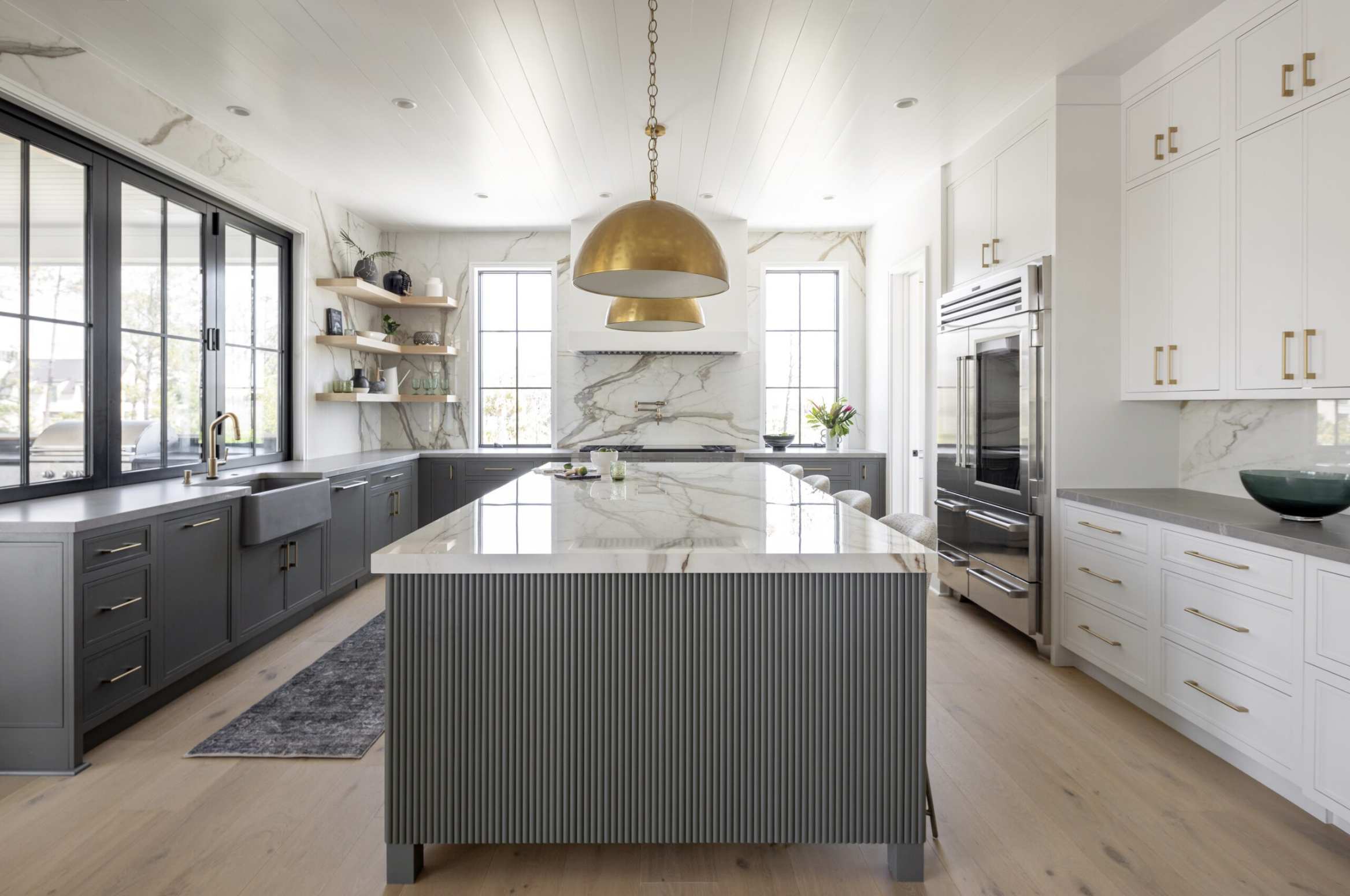 Modern kitchen with white and dark gray cabinetry, marble countertops, large island with striped detail, brass pendant lights, and large windows bringing in natural light.