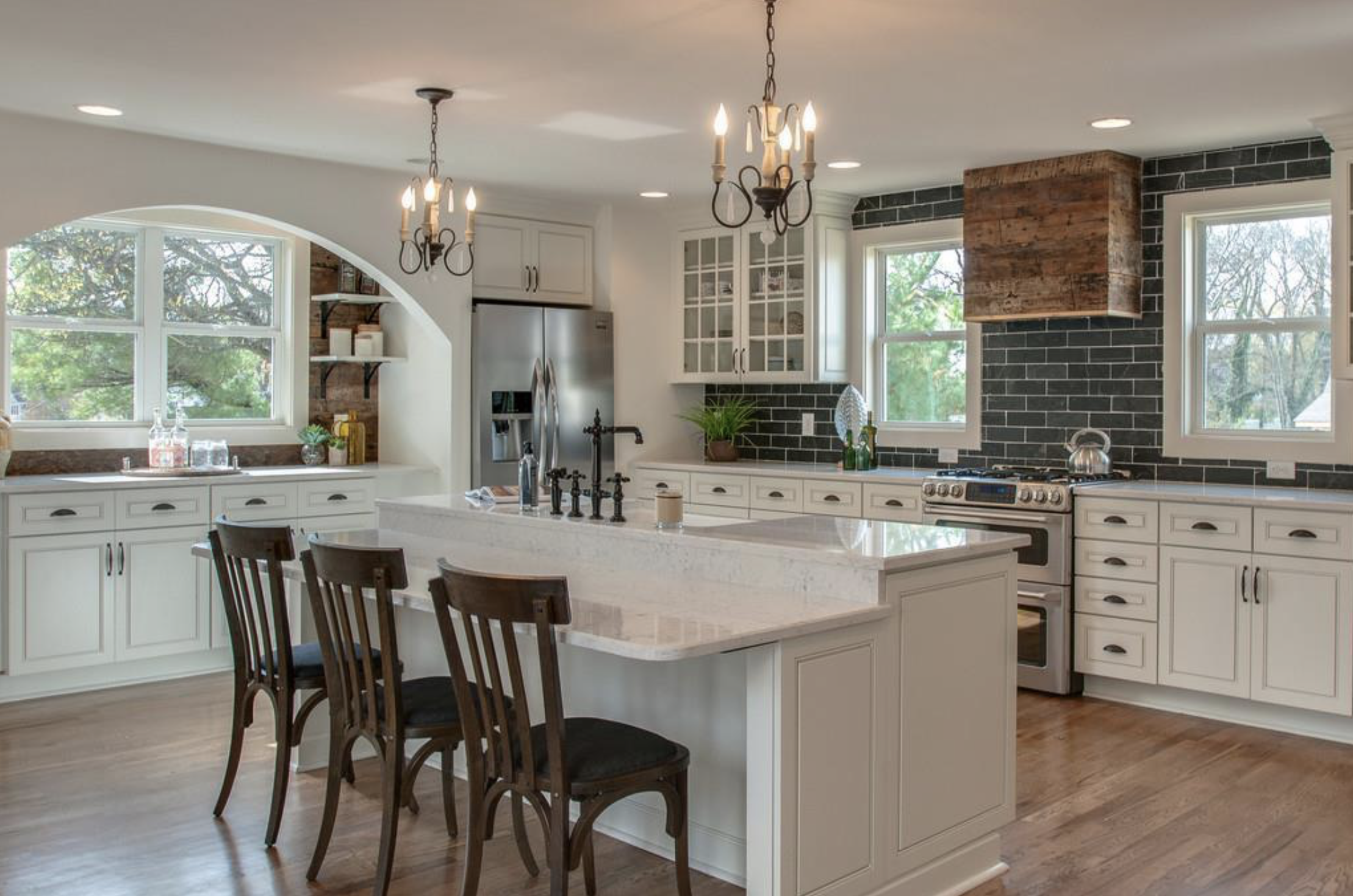 A modern kitchen with white cabinetry, a large island with a light-colored countertop, wooden chairs, black chandelier lighting, and large windows with a view of trees outside. There is a stainless steel refrigerator, a decorative plant, and a chimney feature with wooden panels.