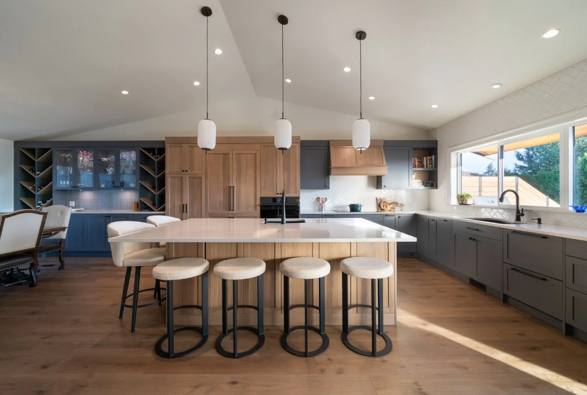 Columbia Cabinets - Modern kitchen with a large island, gray lower cabinets, wooden upper cabinets, and three pendant lights hanging above the island. A window over the sink lets in natural light, and there's a wood floor.