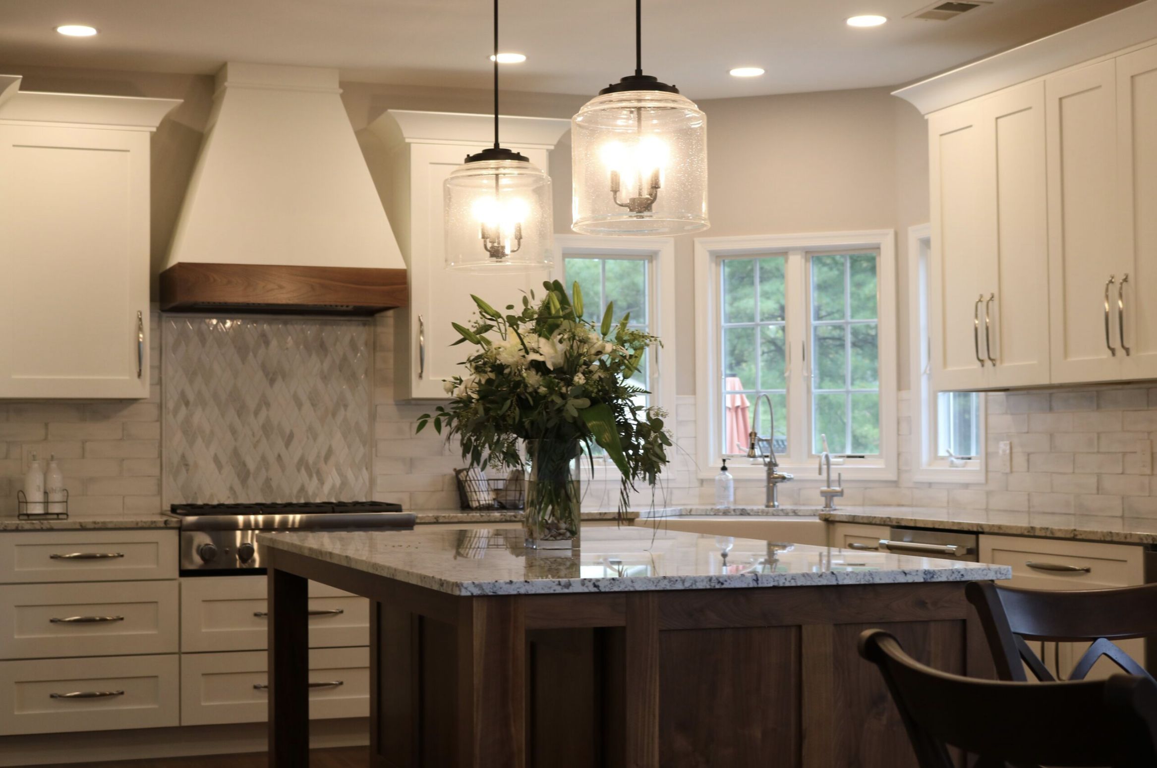 A modern kitchen featuring white cabinets, a large central island with a granite countertop, two pendant lights above, a bouquet of white lilies and greenery on the island, a window behind the sink, and a stove with a wooden range hood.