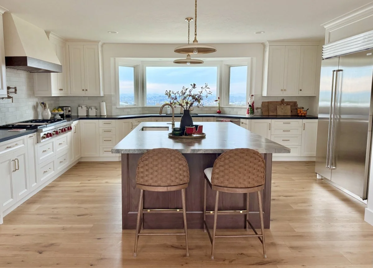 Columbia Cabinets - Bright kitchen with white cabinets, black countertops, a central island with a tray, two woven bar stools, large window with a scenic view, stainless steel refrigerator, and wooden flooring. Kitchen with a view.