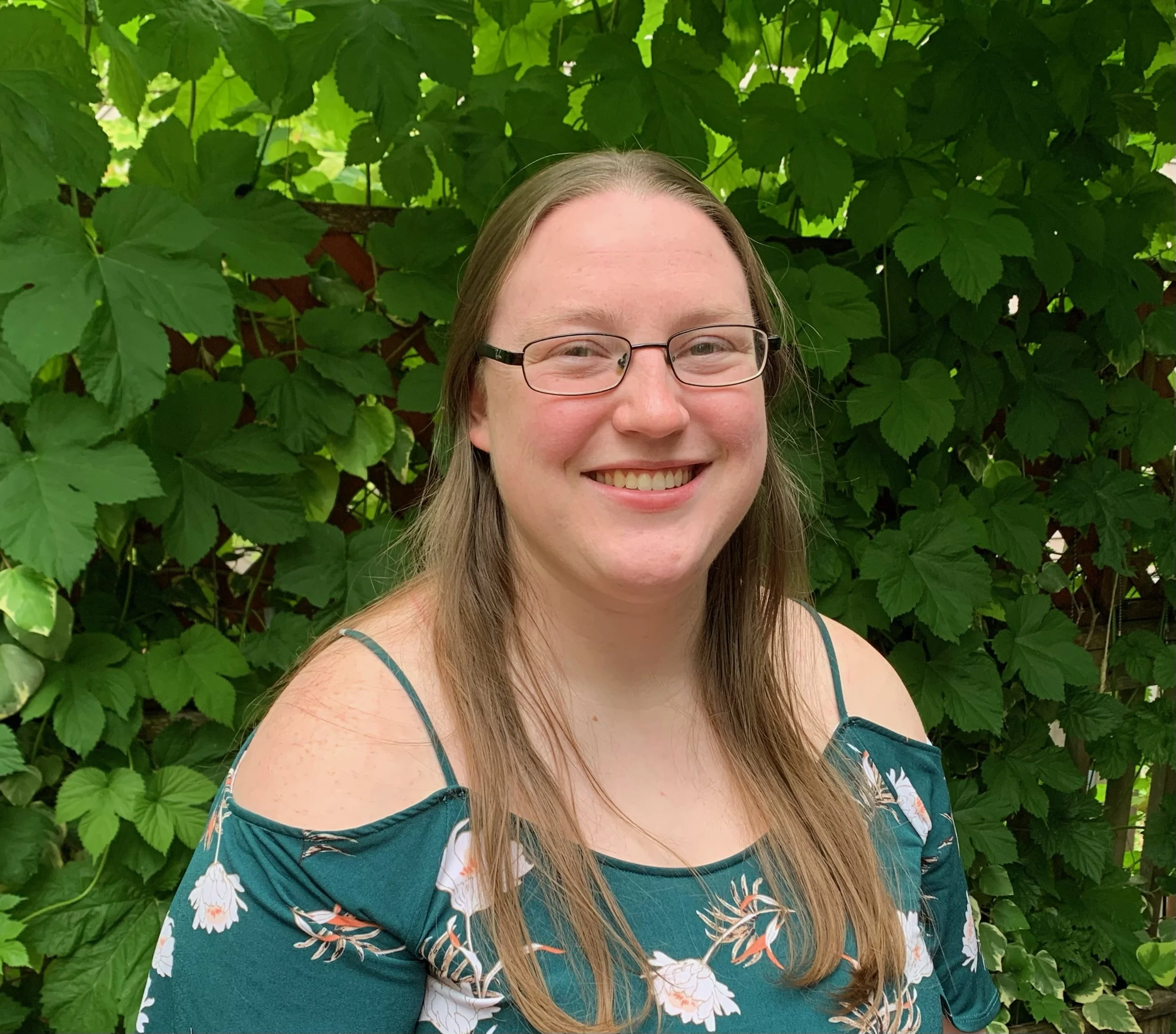 A woman with long brown hair, glasses, and a teal floral dress smiling in front of green leafy plants.