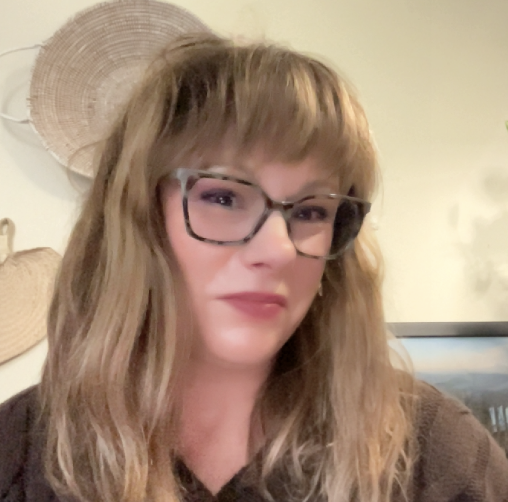 Close-up of a woman with wavy, light brown hair wearing glasses, indoors with wicker decor on the wall.