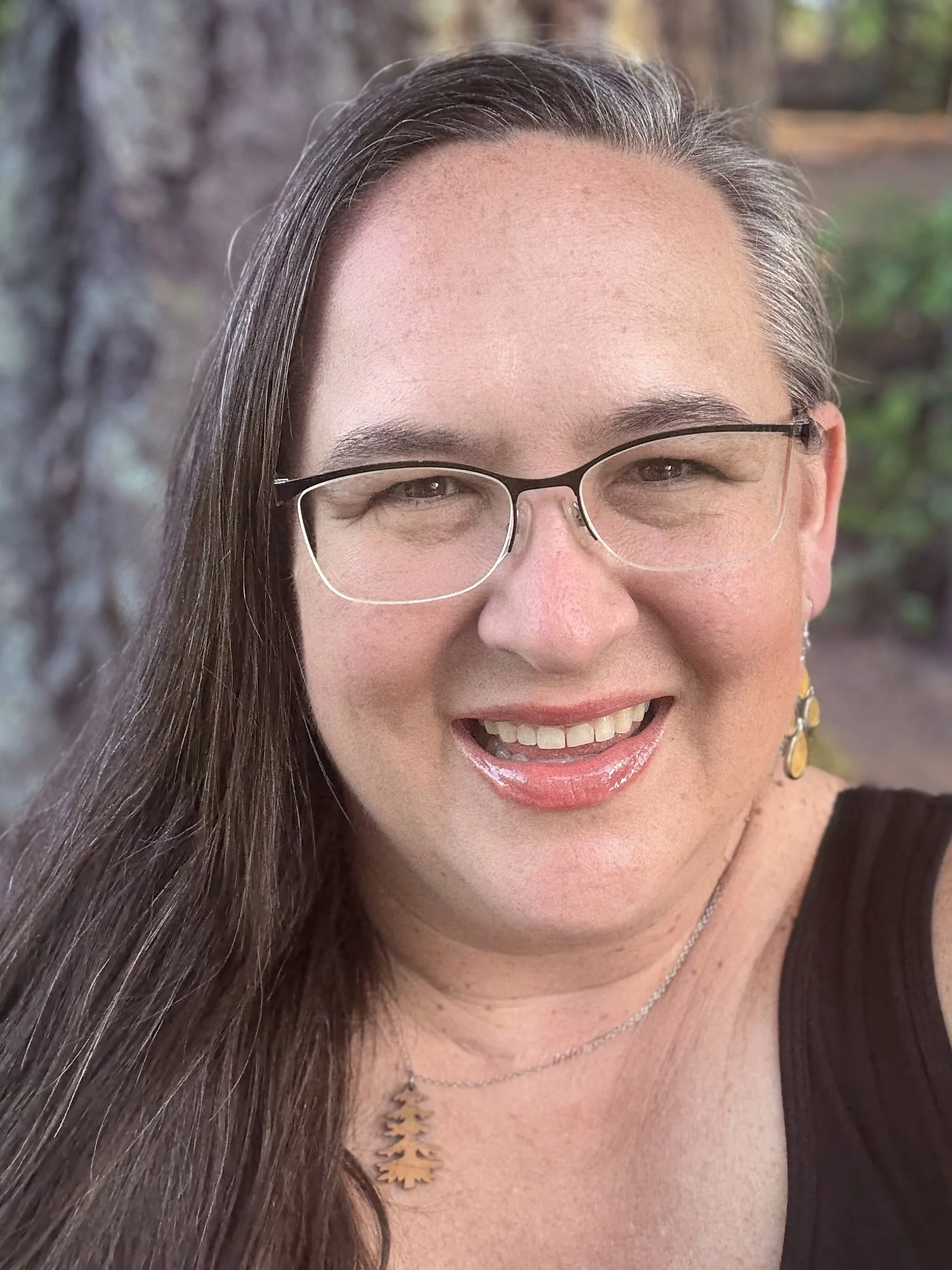 A woman with long brown hair and glasses smiling at the camera outdoors, with trees in the background.