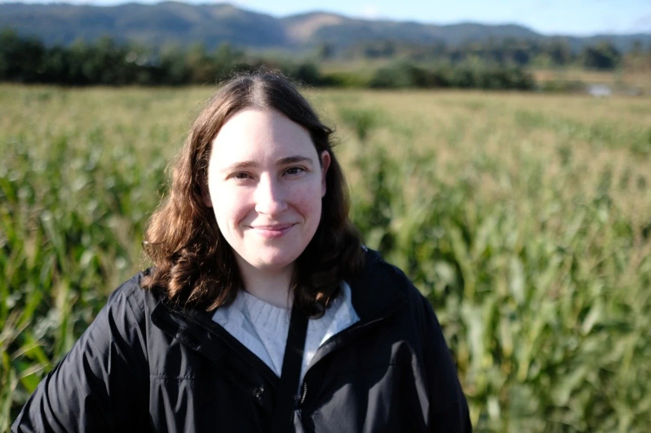 Young woman with brown, wavy hair smiling outdoors in a green field with distant hills in the background.