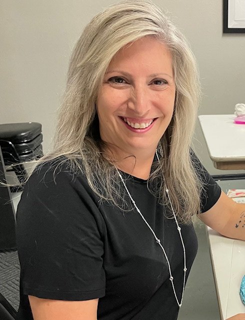 A woman with long blonde hair and a black shirt smiling while sitting at a desk in an office.