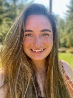 A young woman smiling outdoors on a sunny day, with trees and greenery in the background.