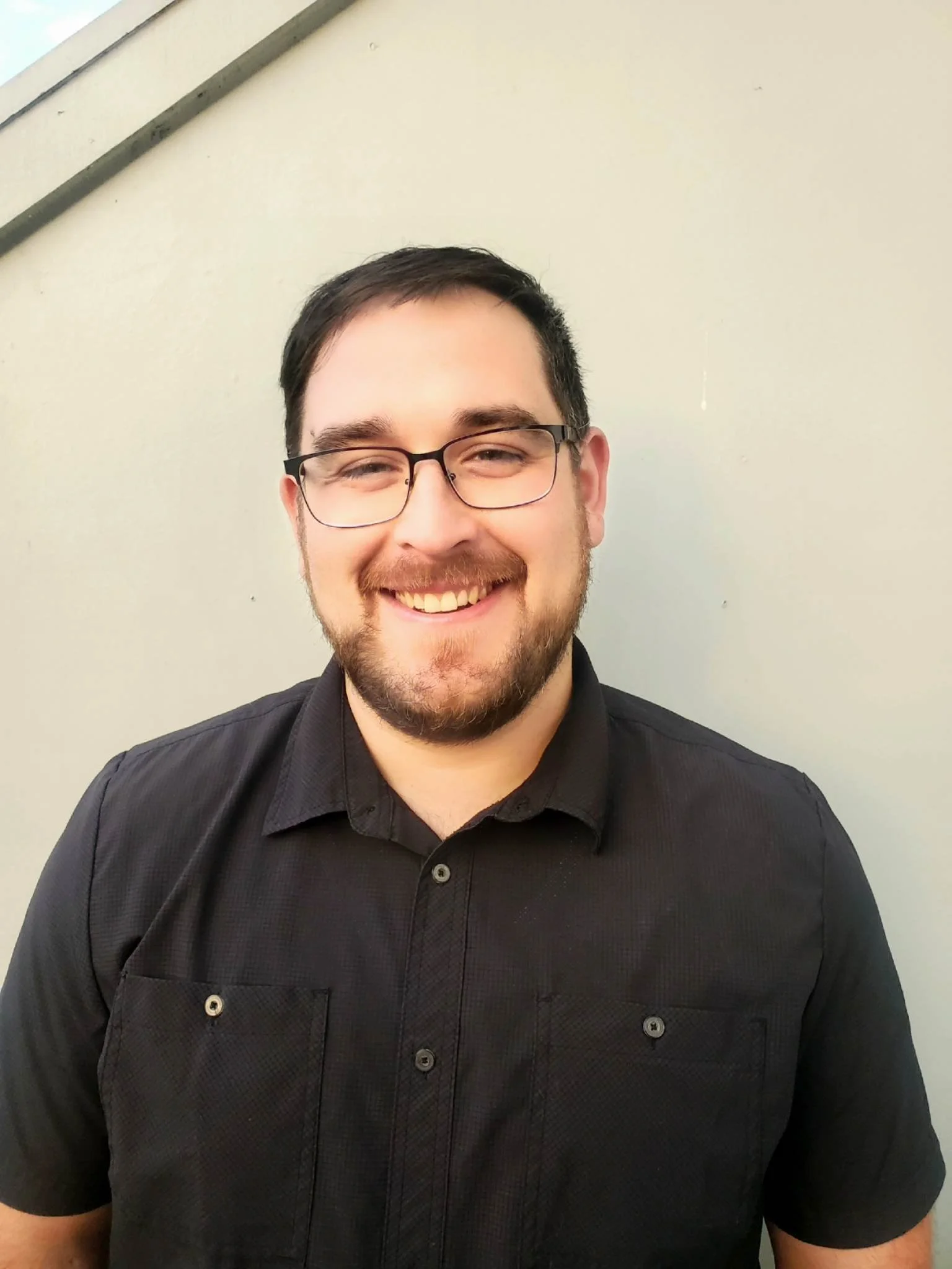 A smiling man with glasses and a beard, wearing a black button-up shirt, standing against a light-colored wall.