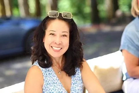Smiling woman with sunglasses on her head, wearing a floral top, outdoors in a park or wooded area.