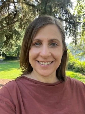 A woman smiling outdoors with trees and greenery in the background.