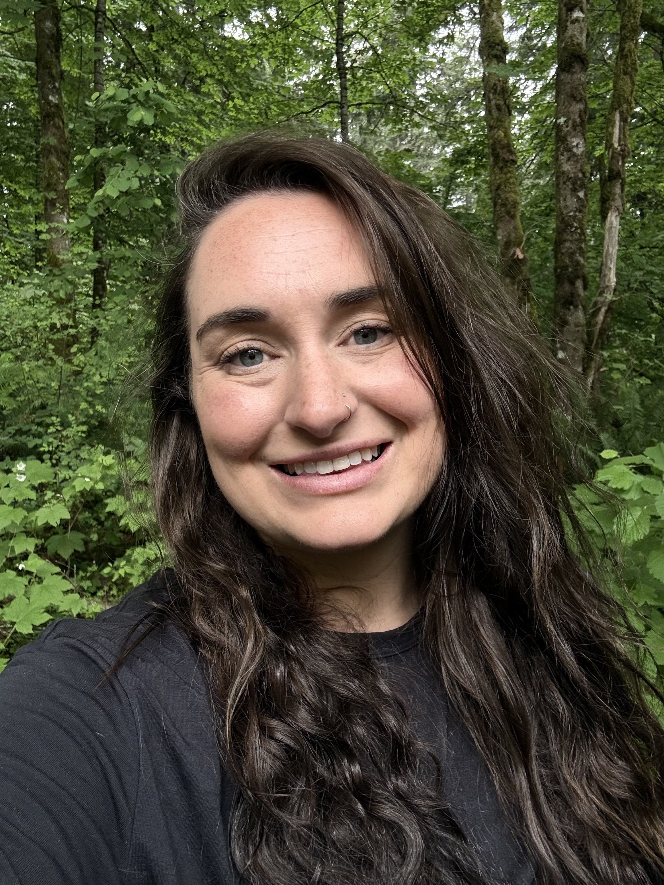 A young woman smiling outdoors on a sunny day, with trees and greenery in the background.