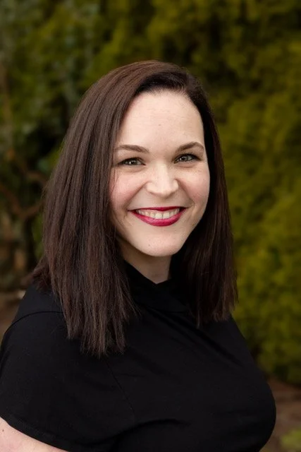 A young woman with long dark brown hair smiling outdoors with a green, blurred background.
