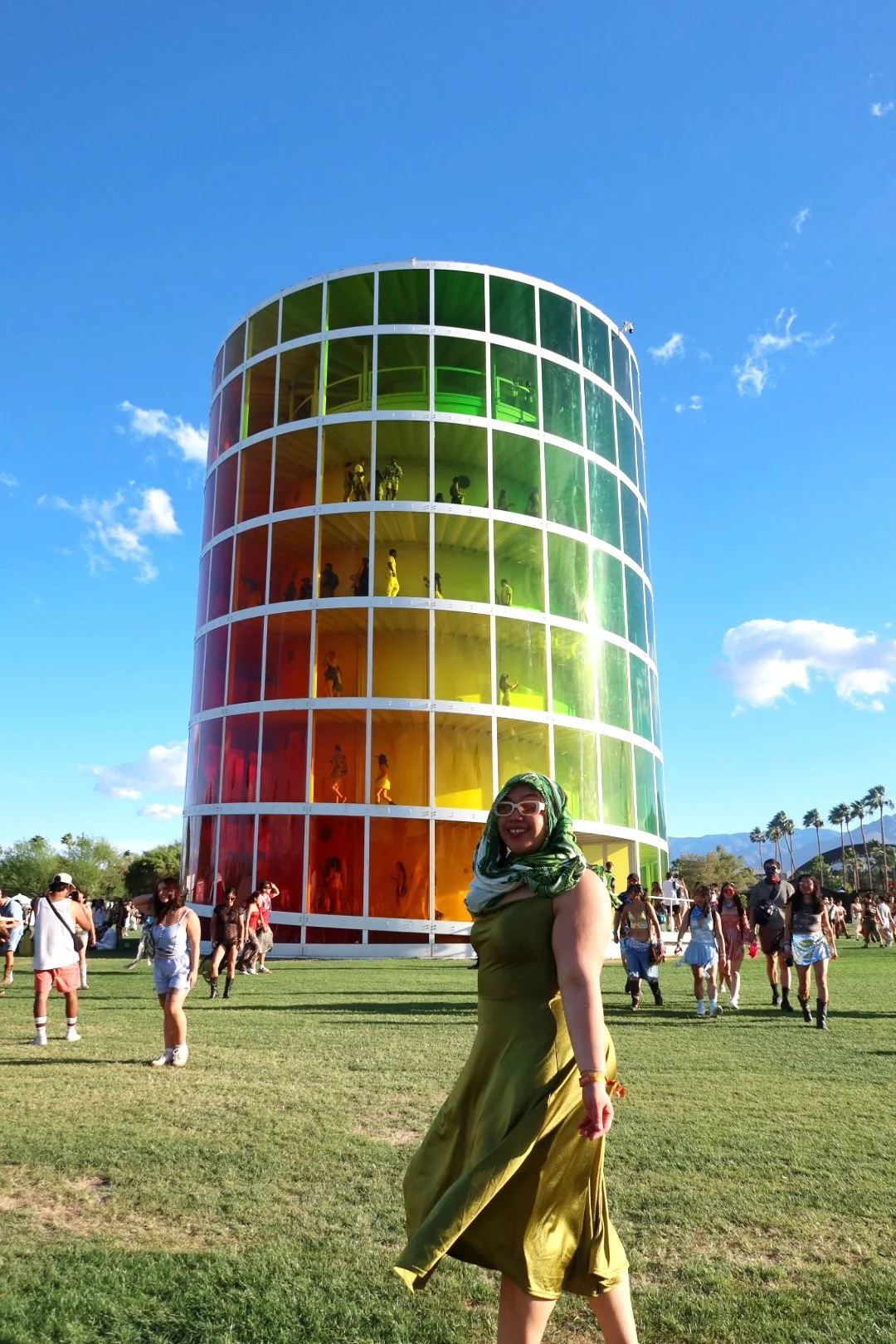 A woman in a green dress and sunglasses smiling at the camera in front of the iCycle, a tall cylindrical building with rainbow-colored glass panels and people visible inside.