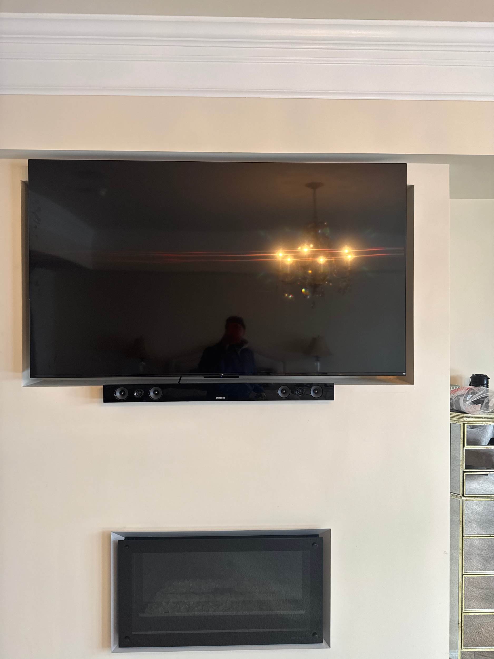 Living room wall with a mounted flat-screen TV and a soundbar below it, along with a wall-mounted heating vent on the right.