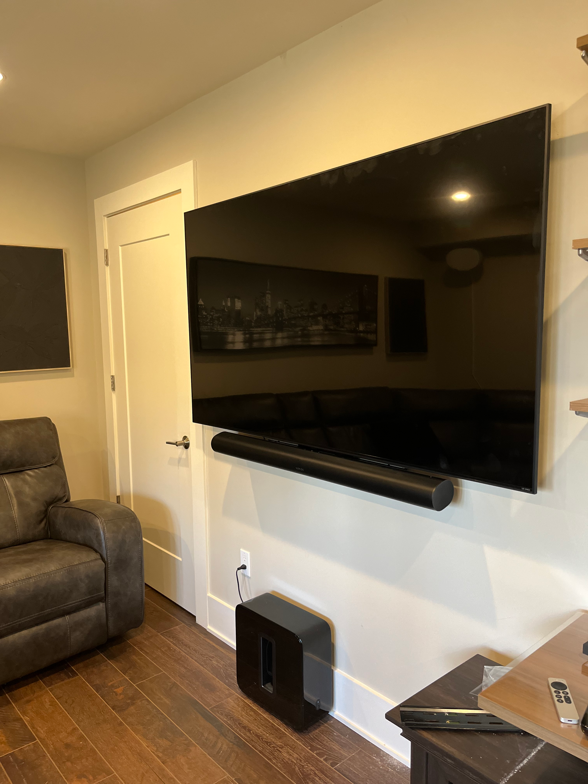 Living room with a mounted flat-screen TV, a soundbar below it, and a leather recliner chair in the foreground. A wooden table with a remote control and electronic device is on the right. The wall is light-colored with a door in the background.