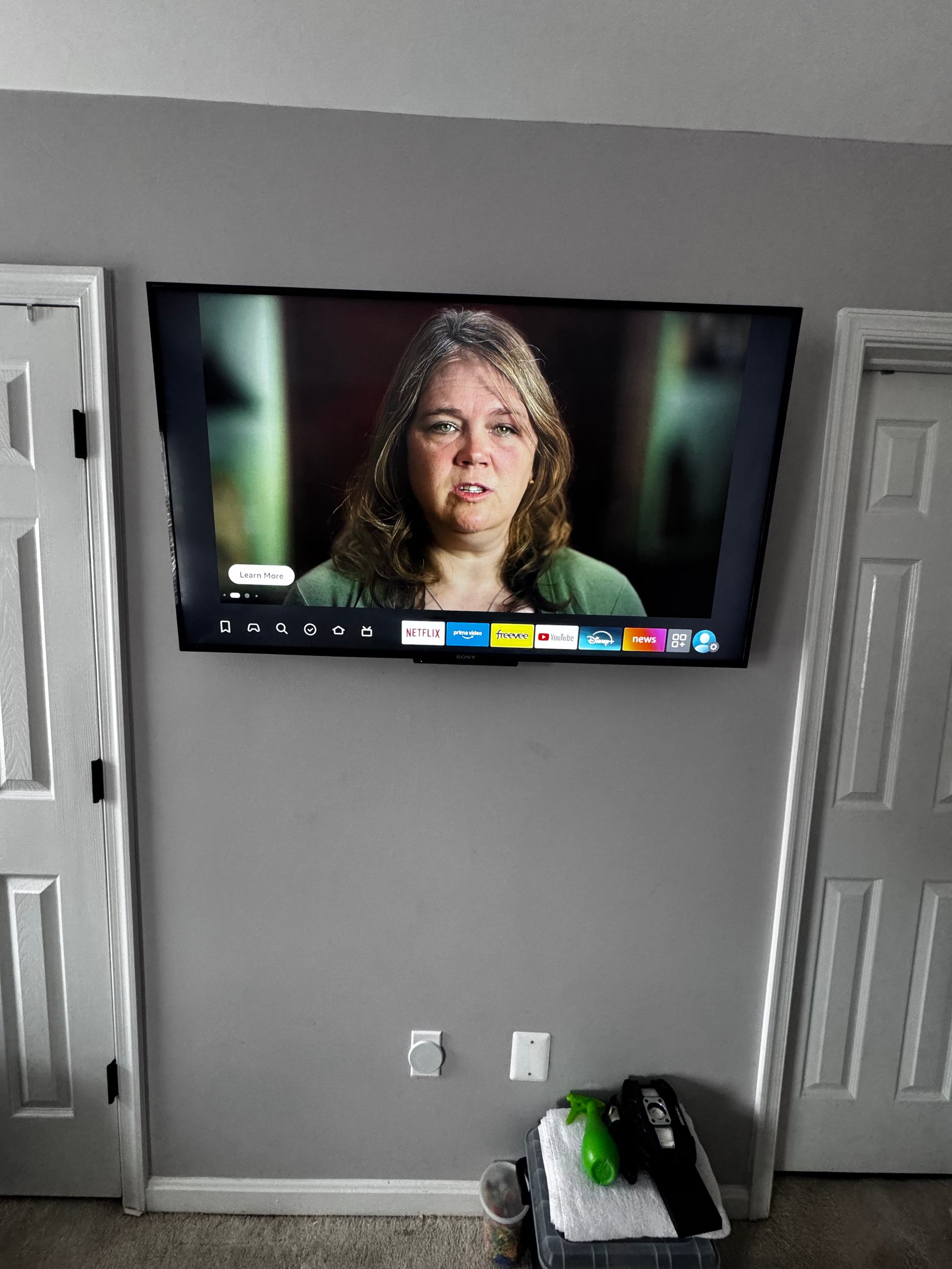 A flat-screen TV on a gray wall displaying a woman with brown, wavy hair and a green top, speaking or talking in a scene from a show or movie.