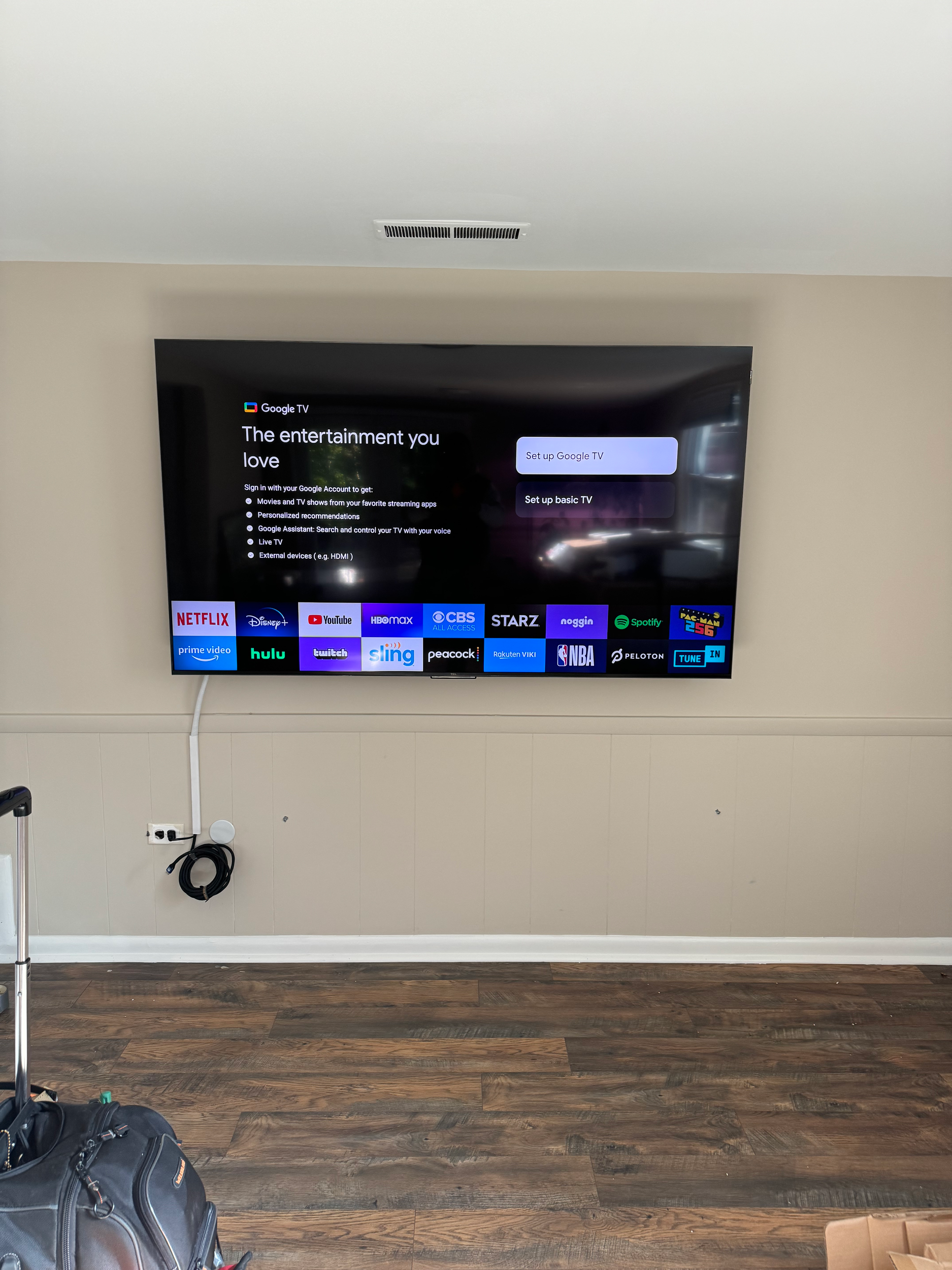 A television mounted on a beige wall displaying the Google TV interface with the message "The entertainment you love" and options for popular streaming services like Netflix, Hulu, and Disney+. A power cord is plugged into an outlet below the TV, and