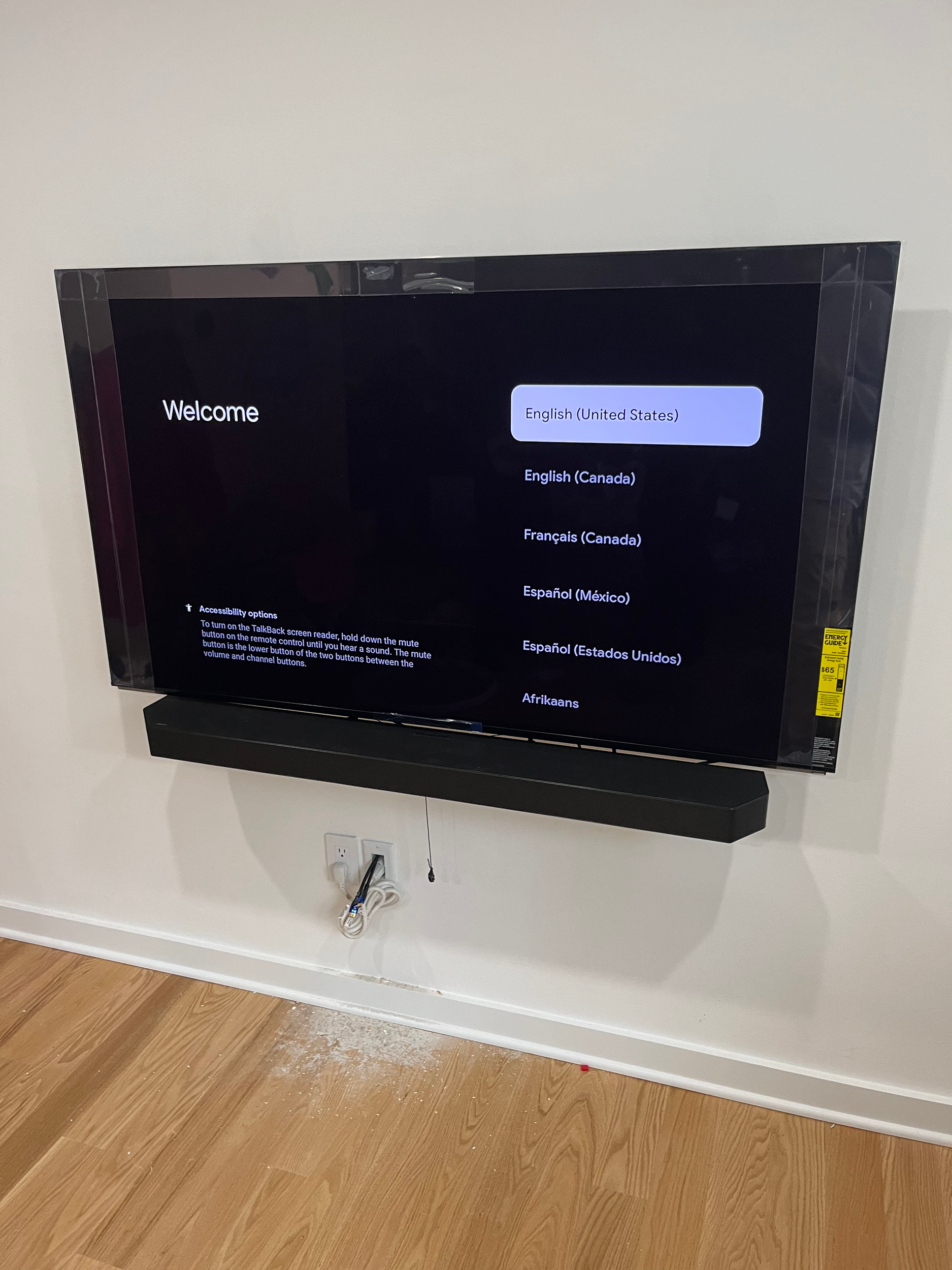 A flat-screen television mounted on a white wall displaying a language selection menu. The menu options include English (United States), English (Canada), Française (Canada), Español (México), Español (Estados Unidos), and Afrikaans. There is a black