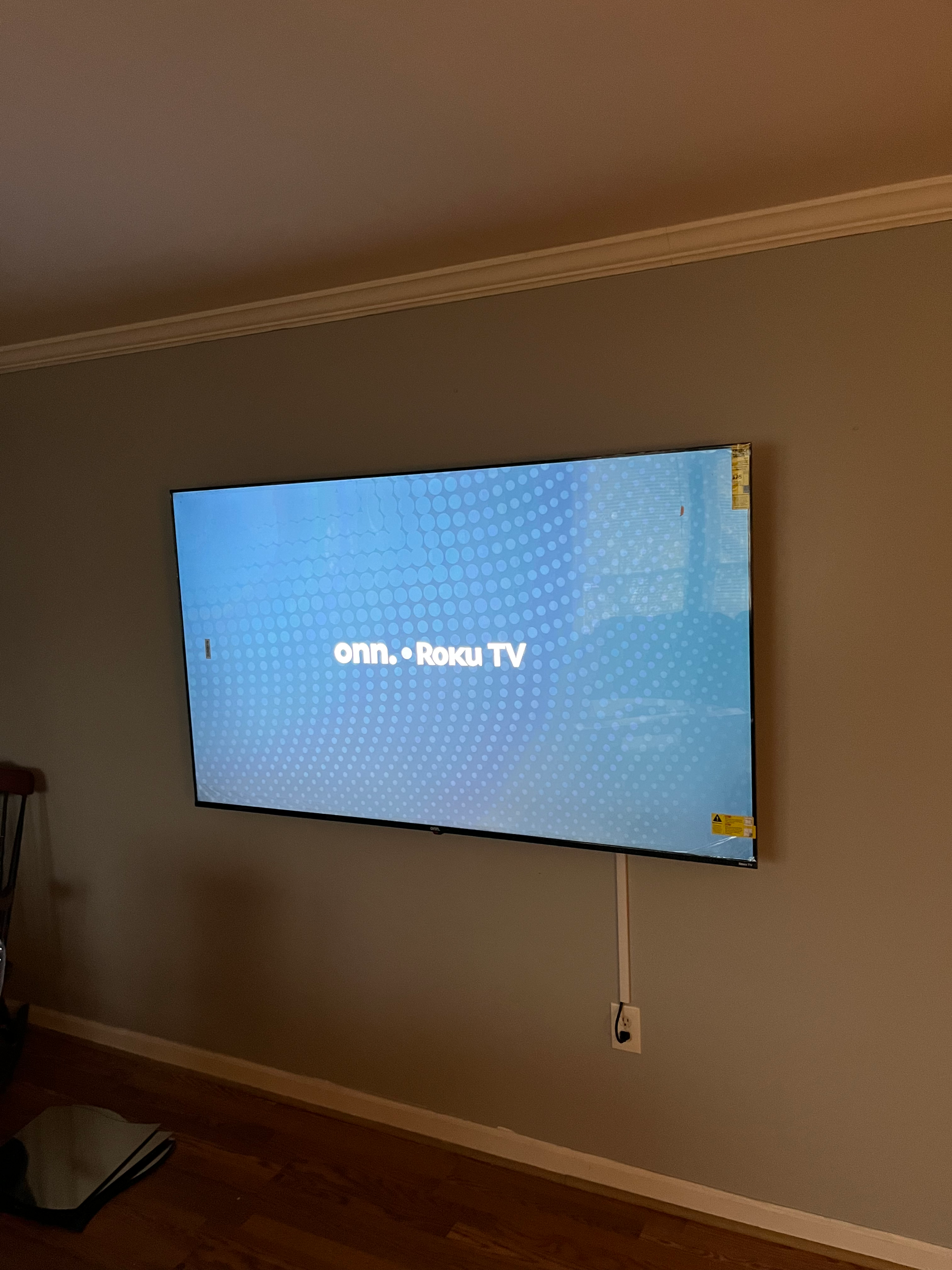 Flat-screen TV mounted on a beige wall displaying the Roku TV startup screen.