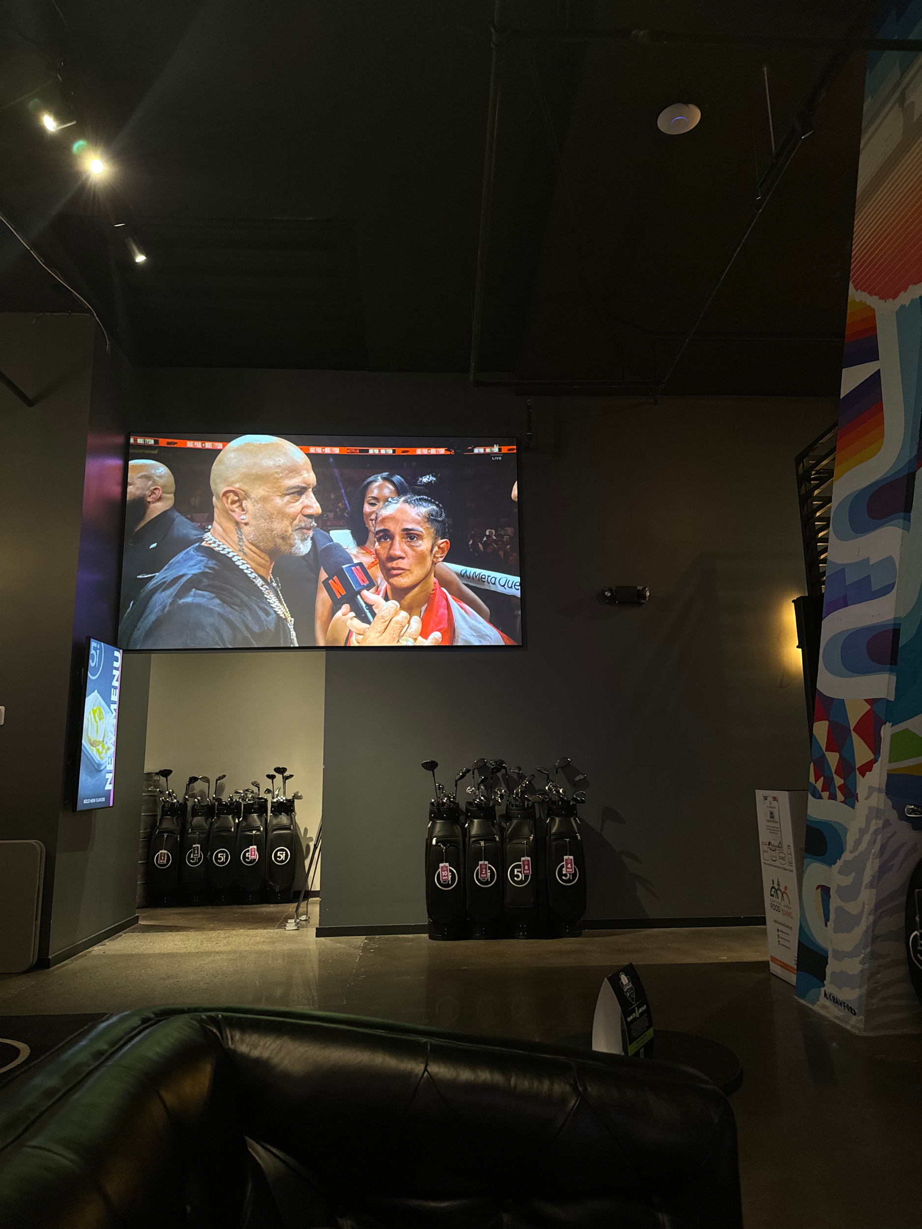 Sports bar or lounge with a large flat-screen TV showing a basketball game, two people sitting on a black leather couch, and a table with multiple speakers on it.