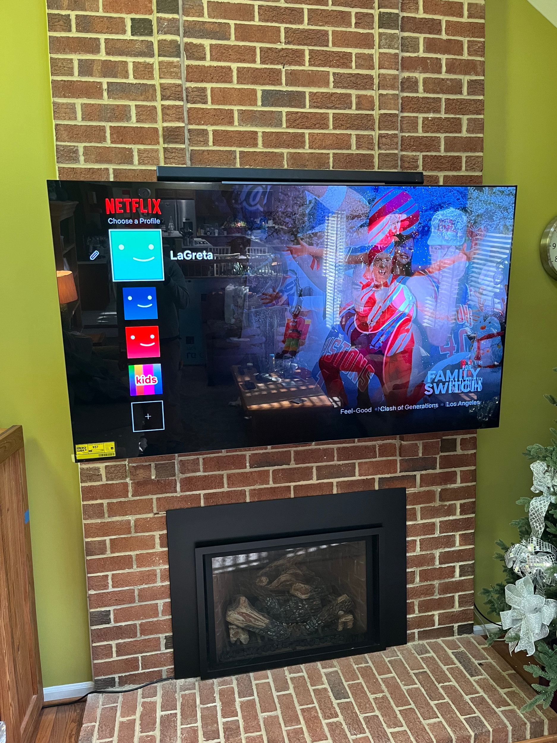 A television screen displaying a Netflix profile selection menu, with a background image of people celebrating at a sports event, mounted on a brick wall next to a fireplace containing logs, with a green wall and a decorative plant nearby.