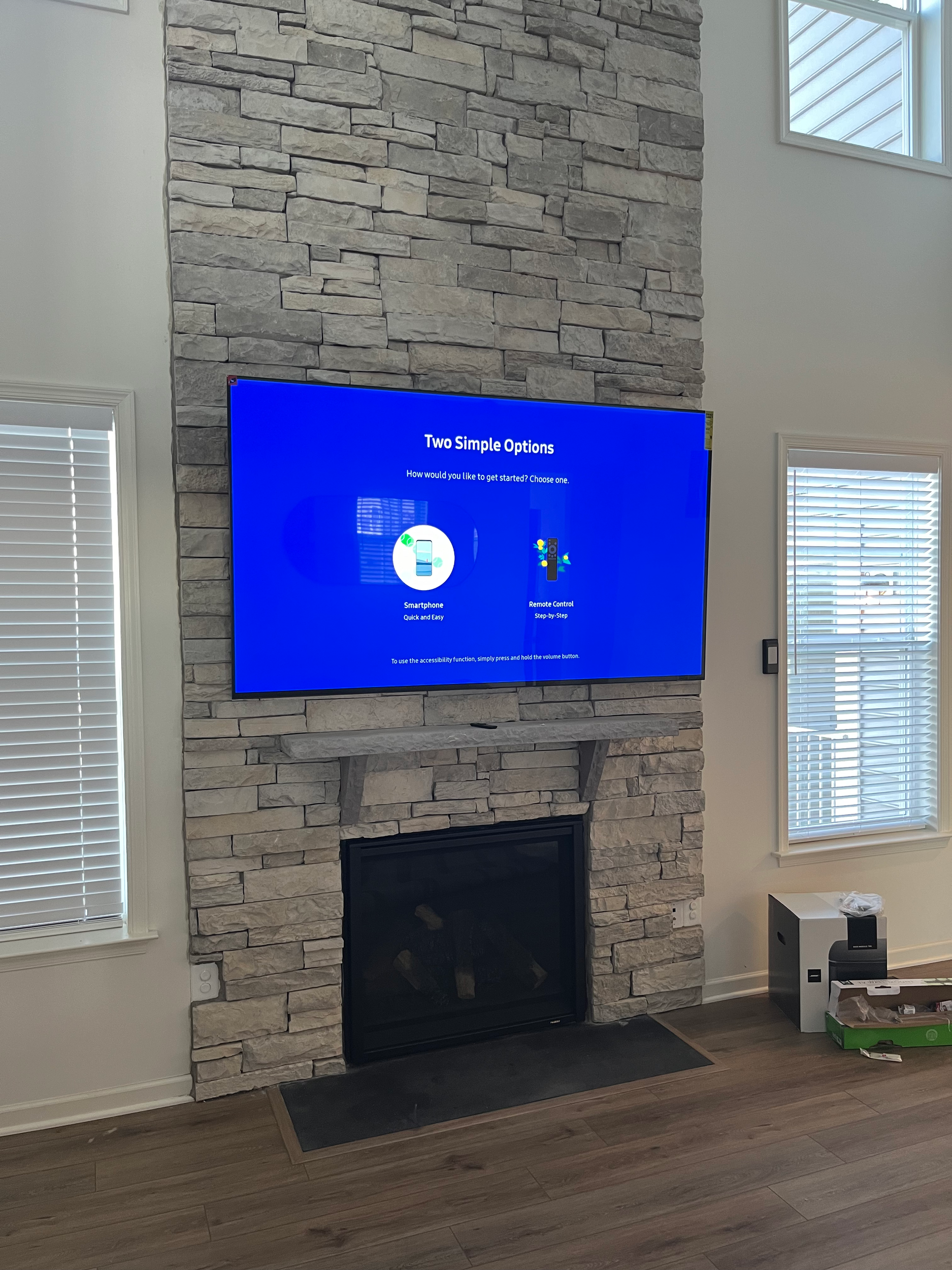 Living room with a stone fireplace and a mounted flat-screen TV on the wall, showing setup options. There are windows with blinds, a small white side table, and a box of tissues on top.