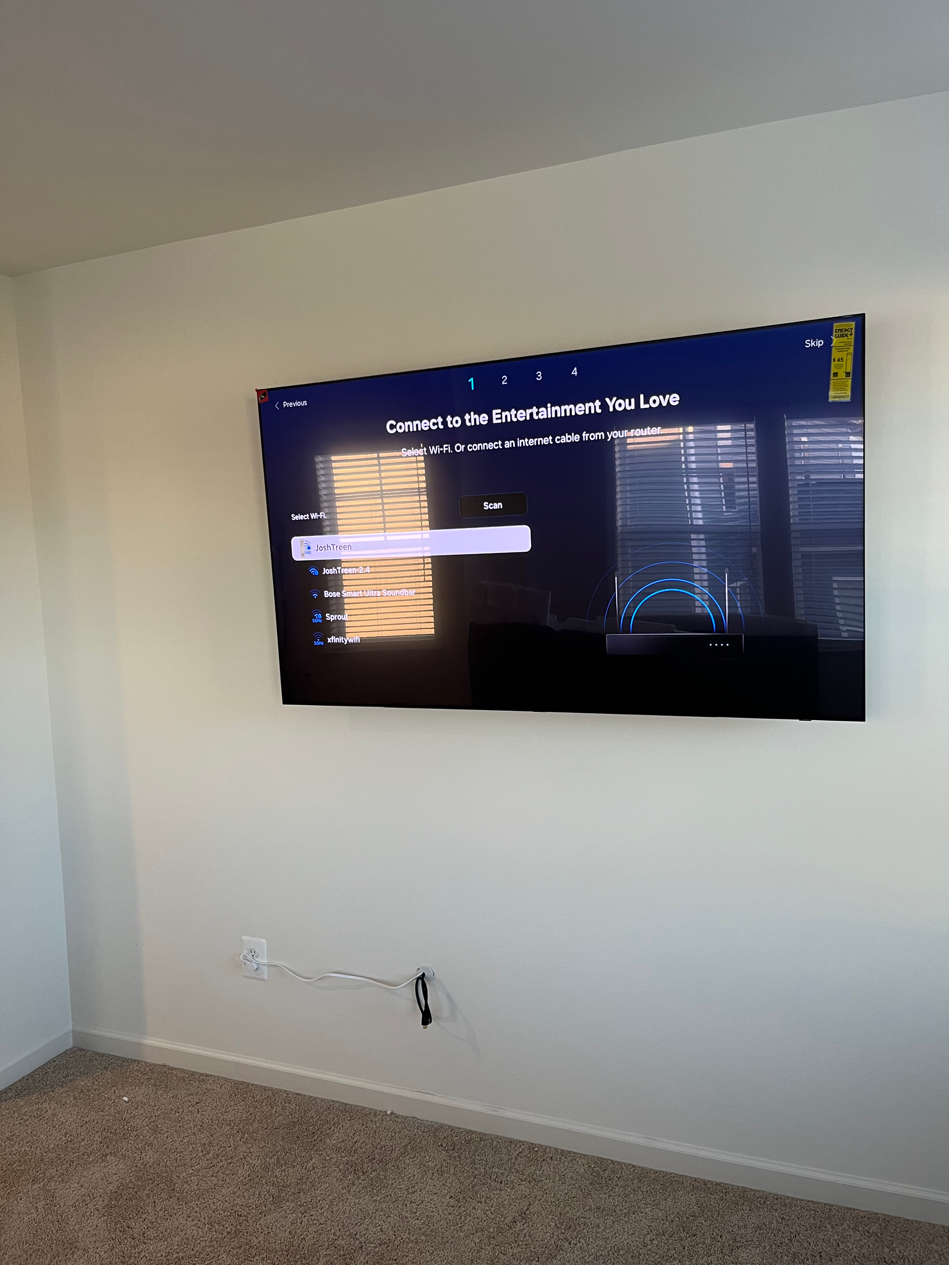 A flat screen television mounted on a white wall displaying a setup screen for connecting to Wi-Fi, with a window with blinds reflected on the screen.