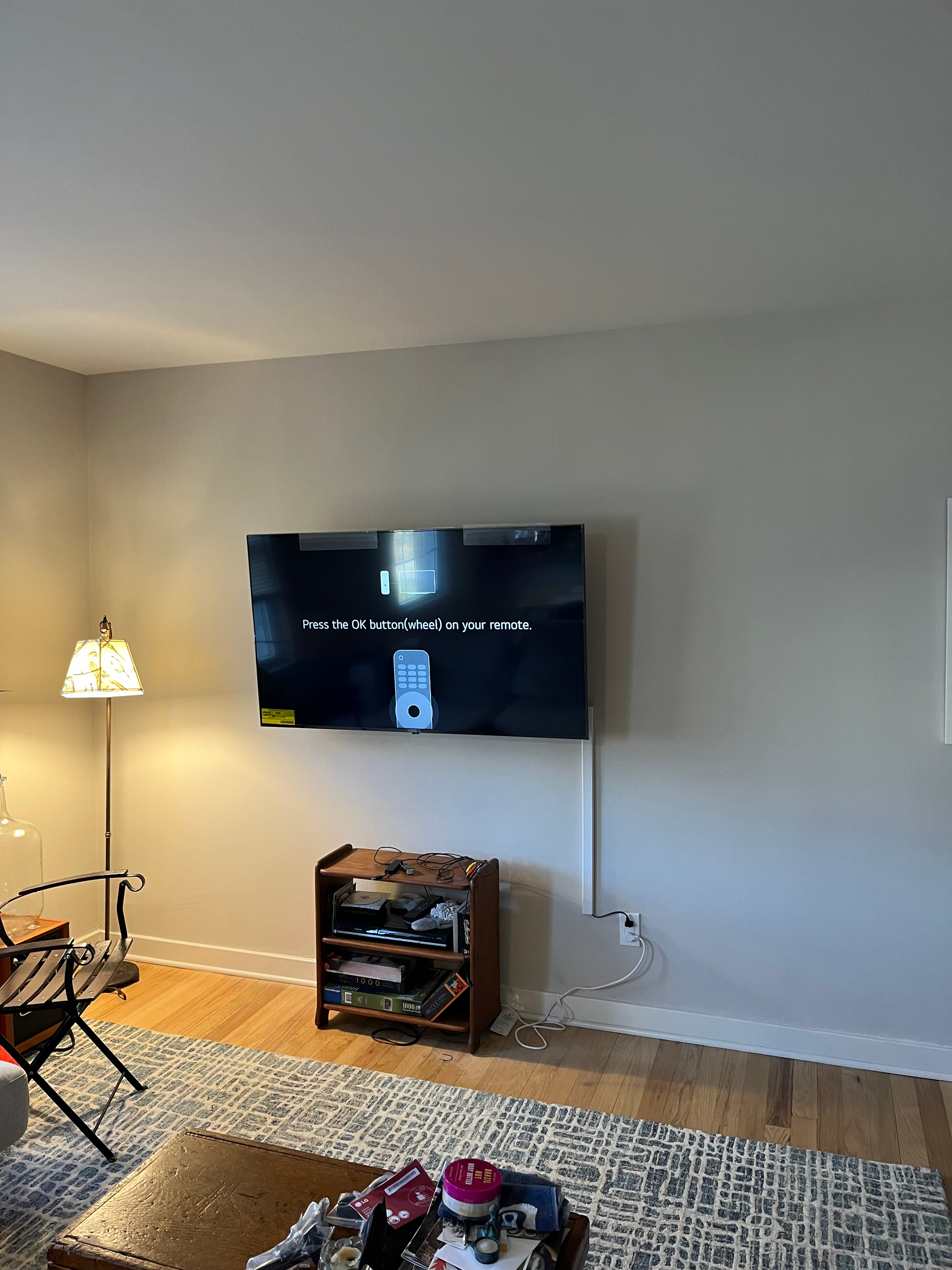 Living room with a mounted flat-screen TV, entertainment stand with electronic devices, a standing lamp, a patterned rug, and some clutter on a coffee table.