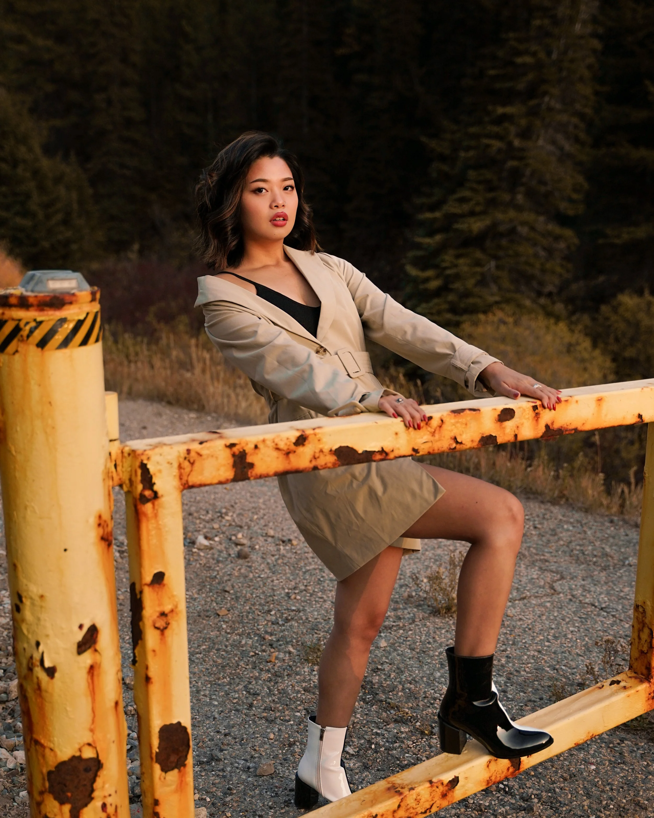 A woman in a beige trench coat and black top standing behind a rusty yellow metal barrier outdoors with trees in the background.