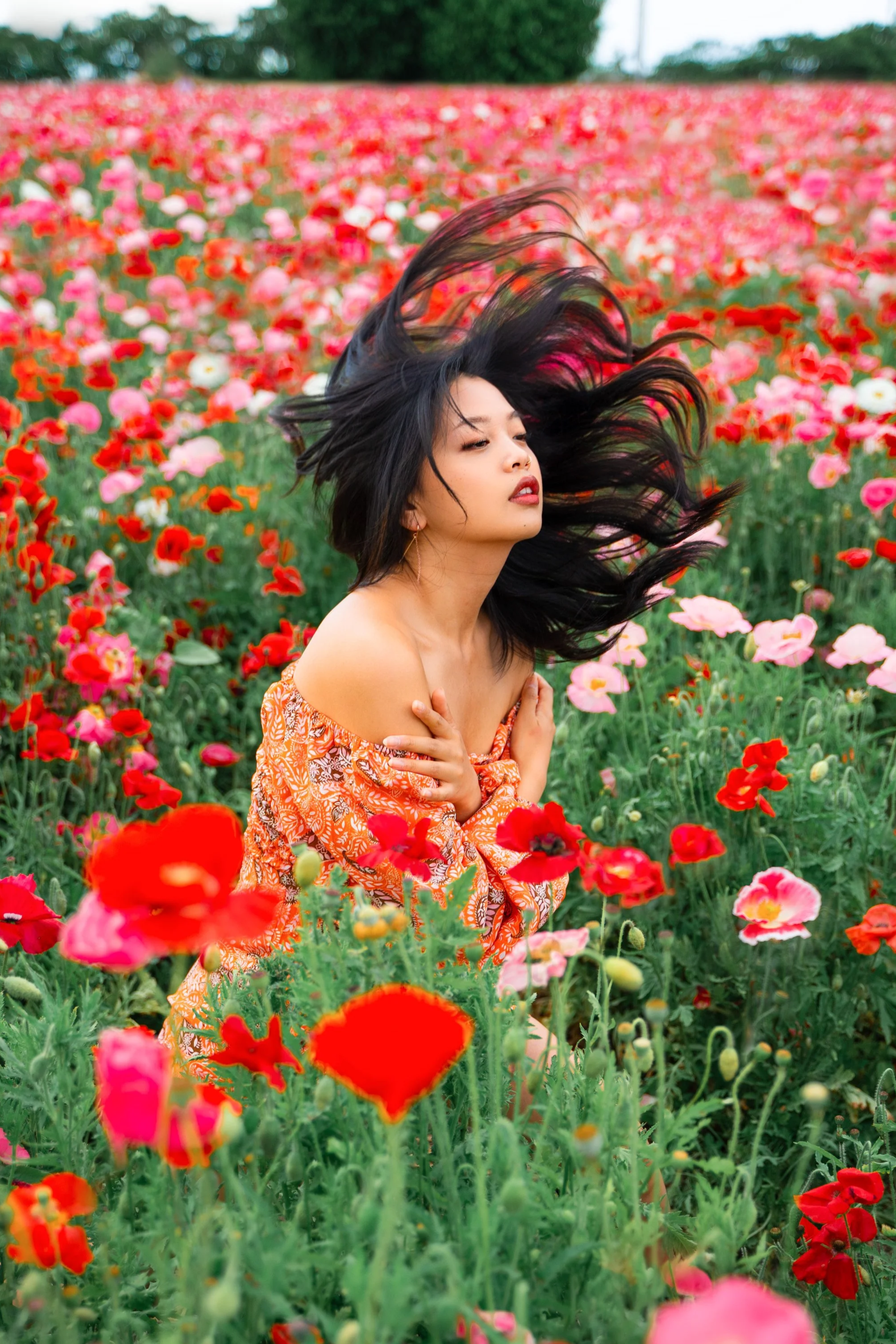A woman with long black hair and a patterned orange dress standing in a field of pink and red poppies, with trees and a blue sky in the background.