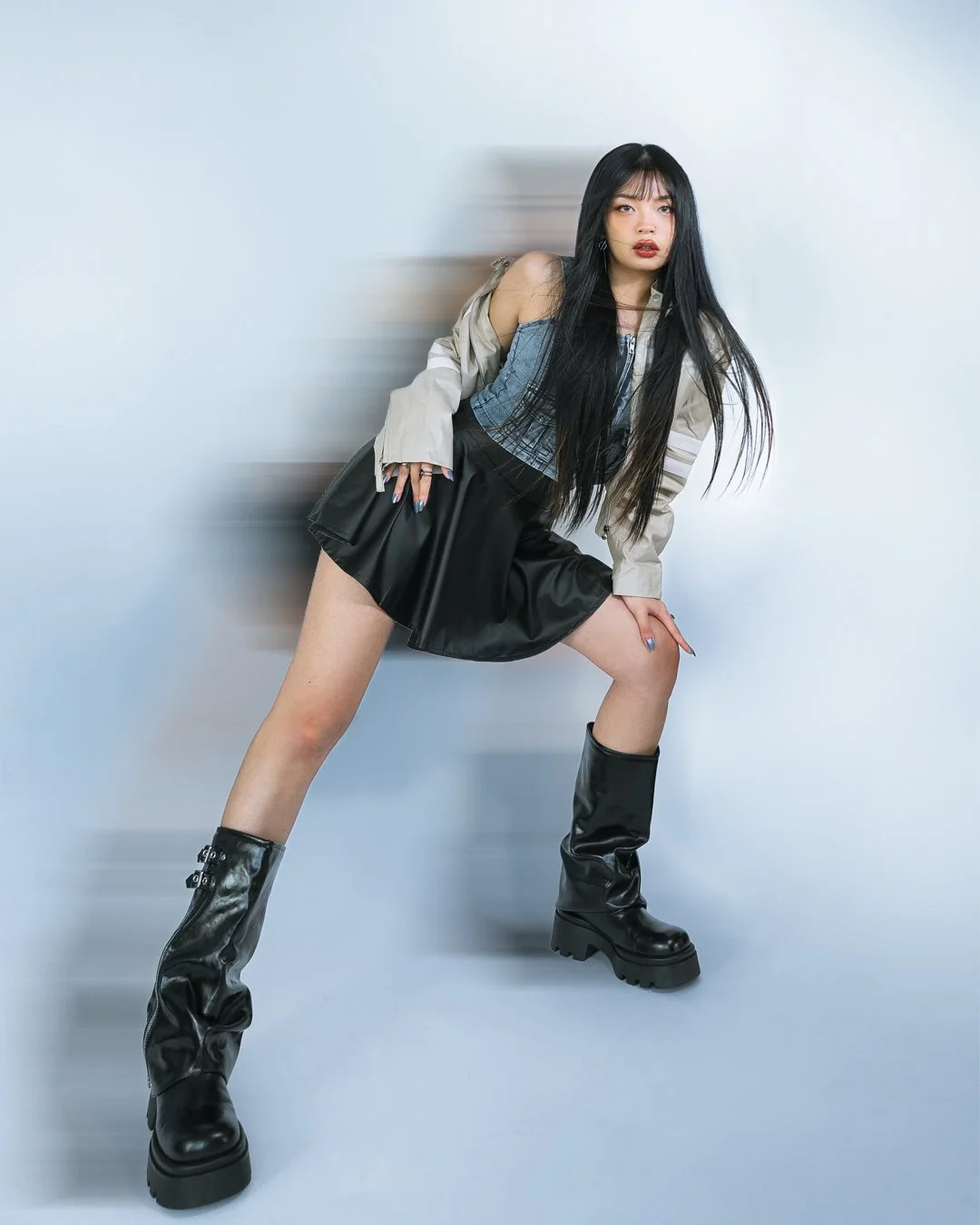 A young woman with long black hair poses against a plain wall. She is wearing a beige jacket, a blue top, a black skirt, and black chunky knee-high boots. She has a confident expression and is leaning with one hand on her thigh.