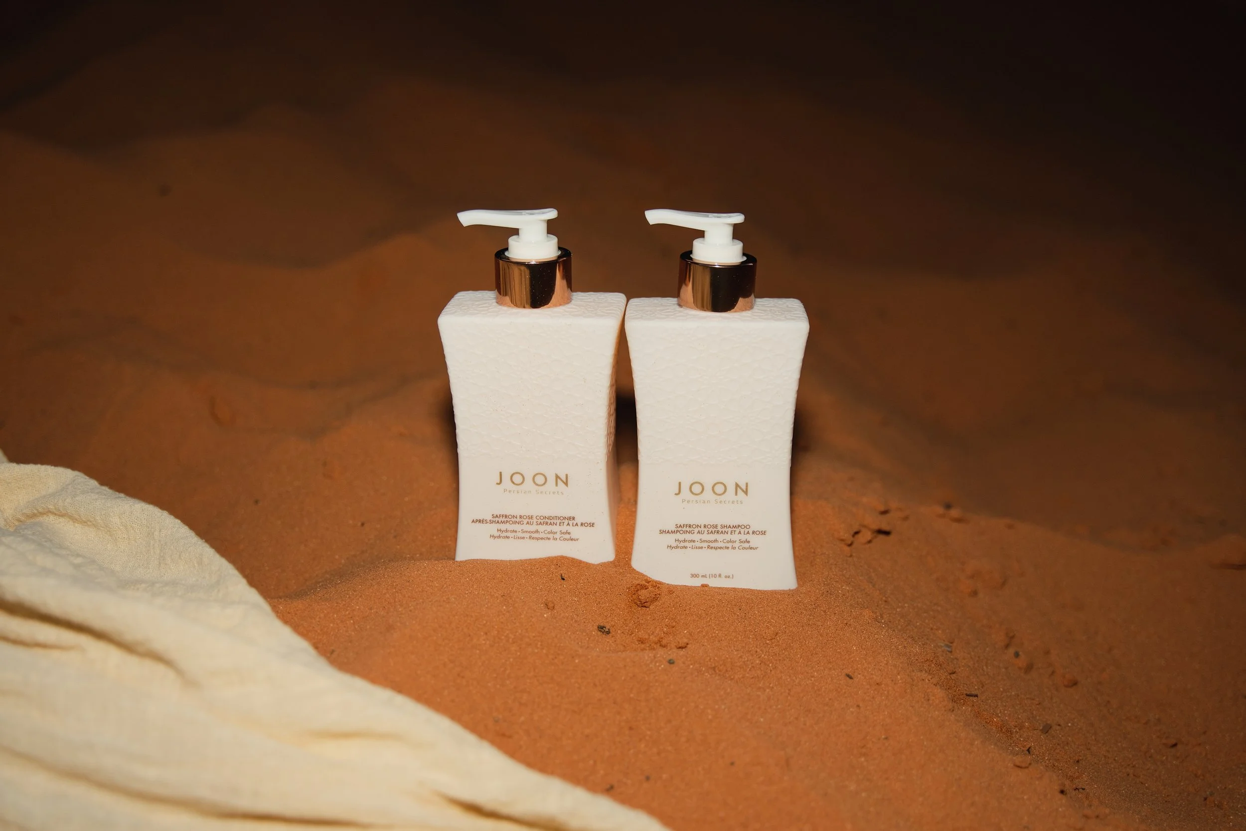 Two bottles of JOON Persian Secrets hair care products, Saffron Rose Conditioner and Saffron Rose Shampoo, placed on sandy ground with a beige cloth nearby.