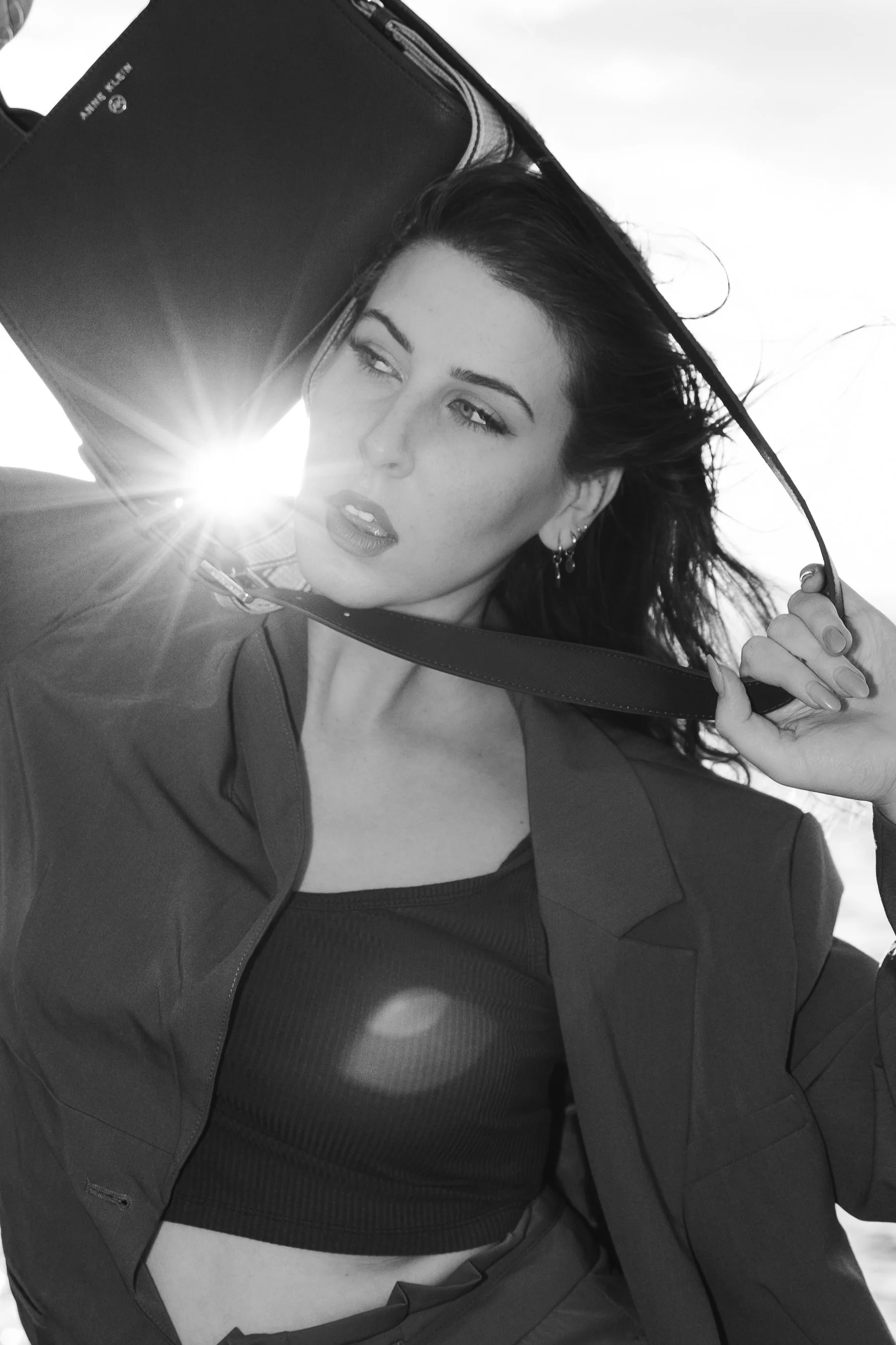A woman with dark hair, wearing a jacket and a black sports bra, holds a large black umbrella. The sun is shining behind her, creating a starburst effect.