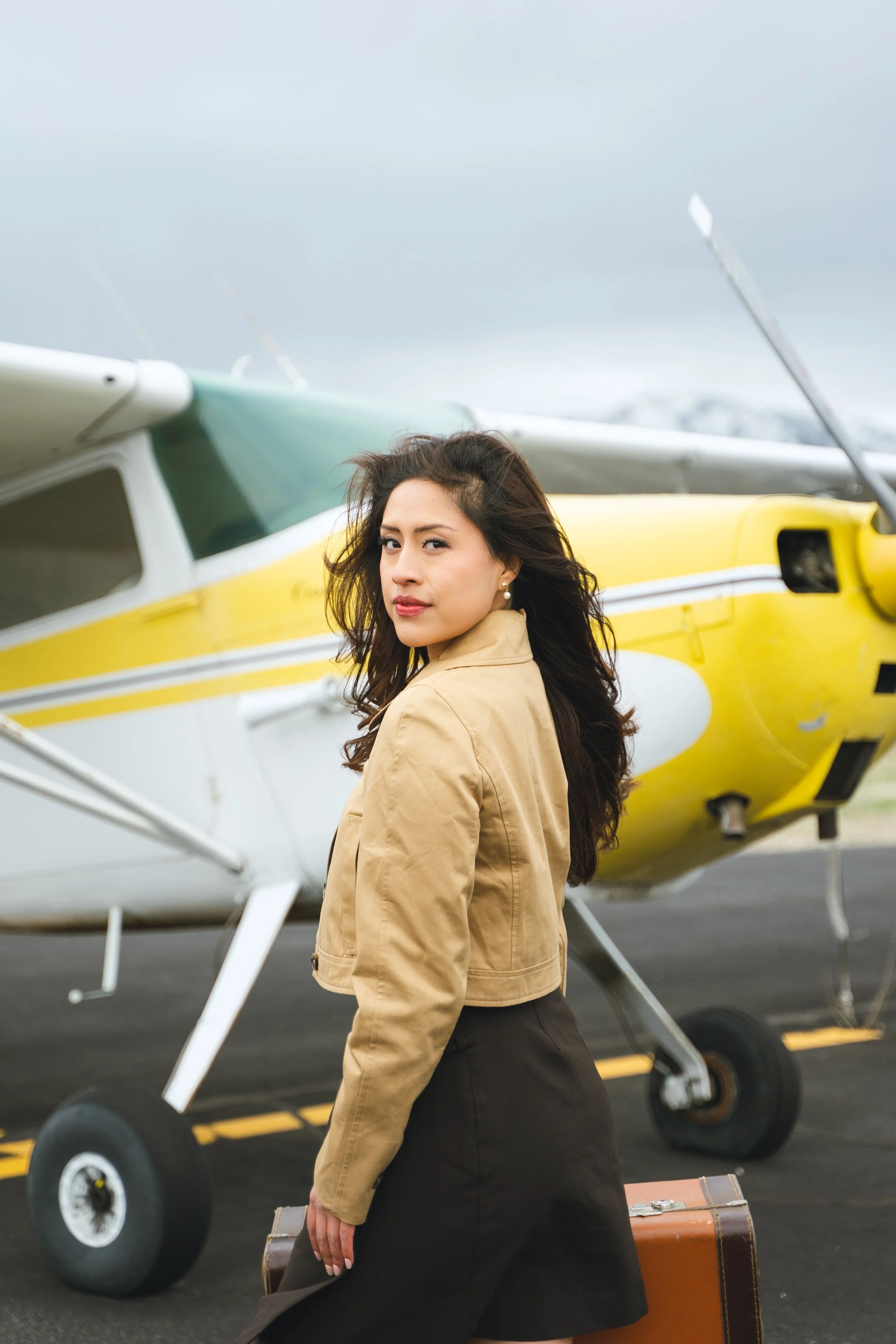 A woman with long dark hair, wearing a beige jacket and black skirt, stands in front of a yellow and white helicopter on an airport tarmac. She is holding a brown leather suitcase and appears to be ready to travel.