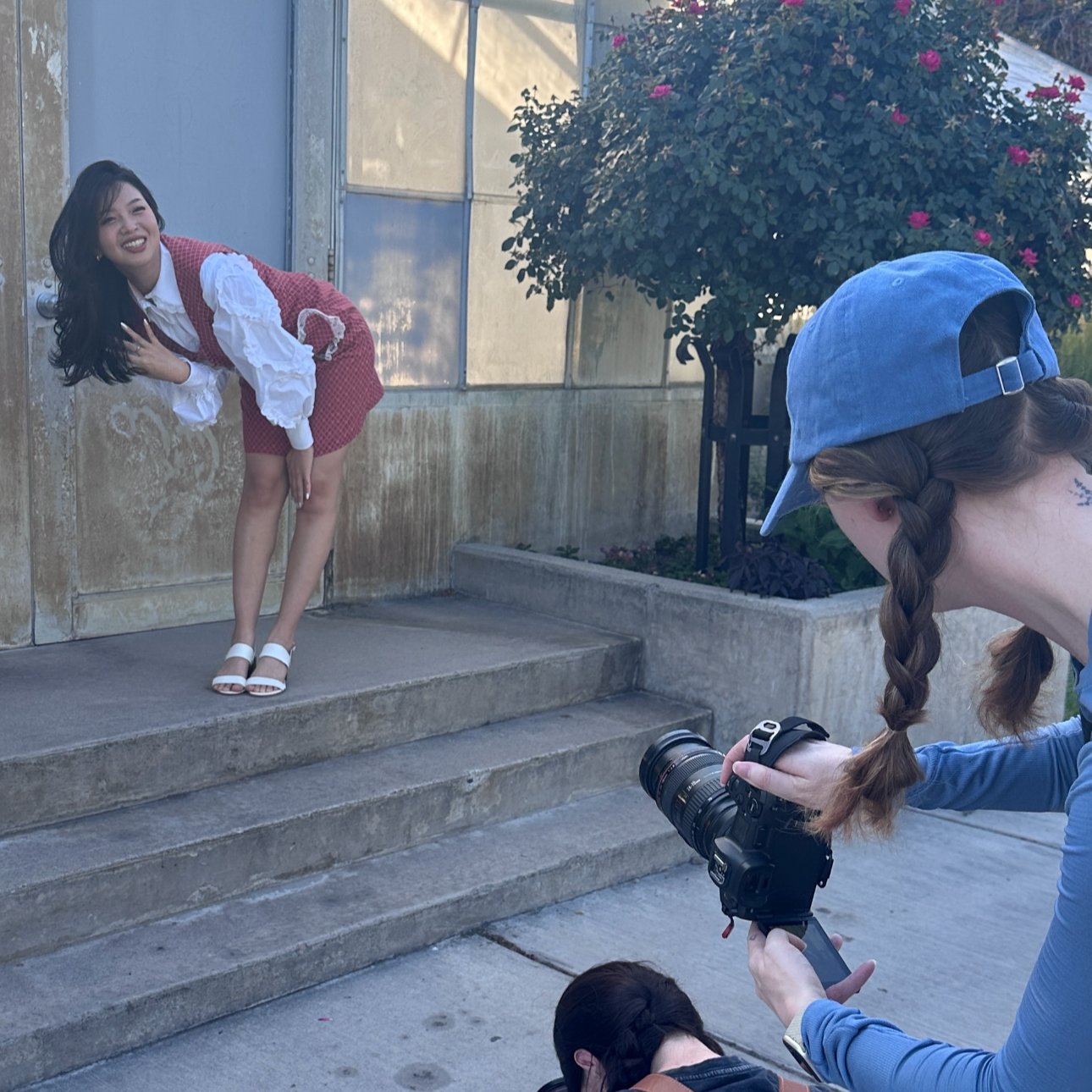 A woman in a red checkered dress with white blouse and white sandals is posing on concrete stairs, leaning sideways and smiling, while a photographer with braided hair, blue cap, and camera takes her picture outdoors near a flowering shrub.