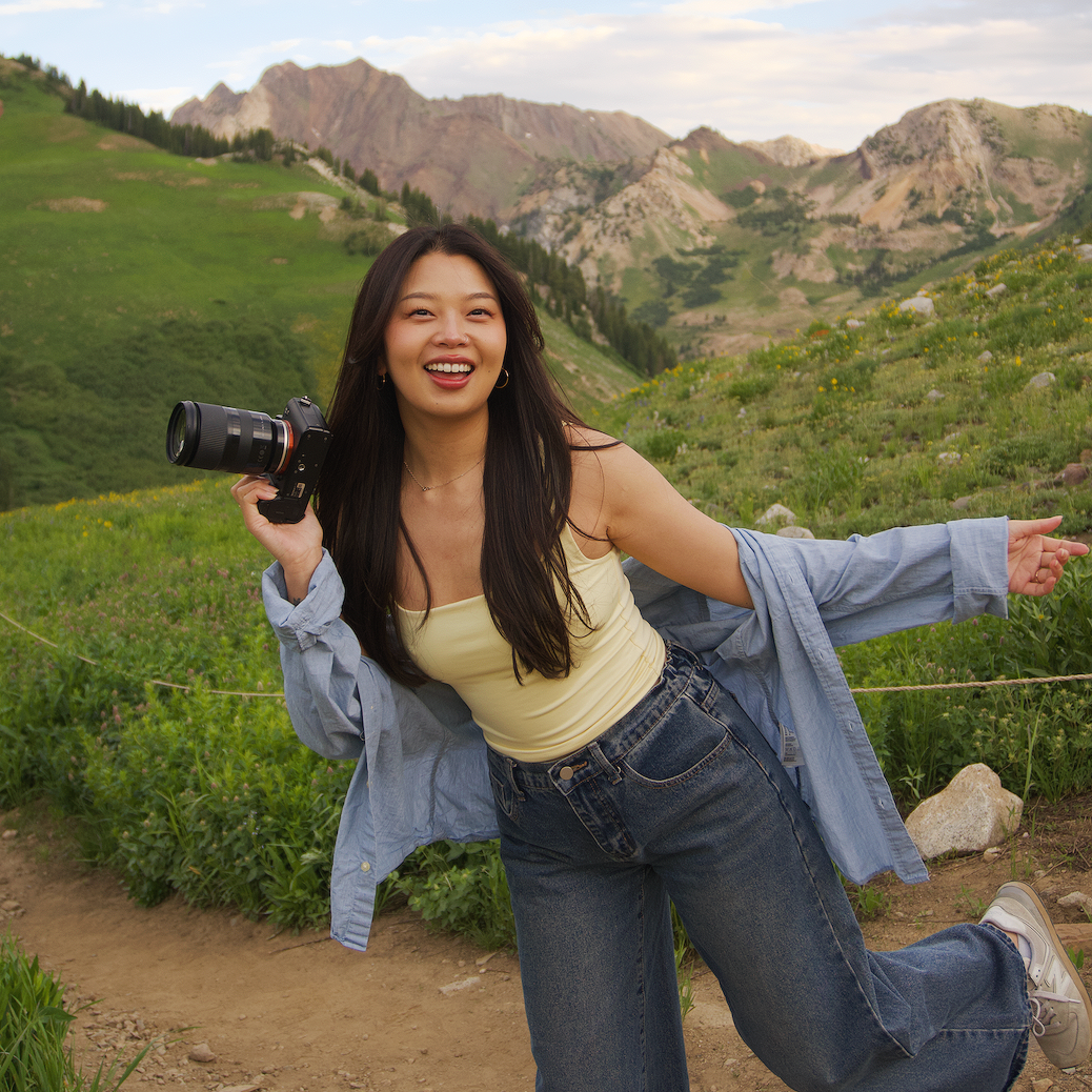A smiling woman with long dark hair holding a camera in an outdoor landscape of green hills and mountains.
