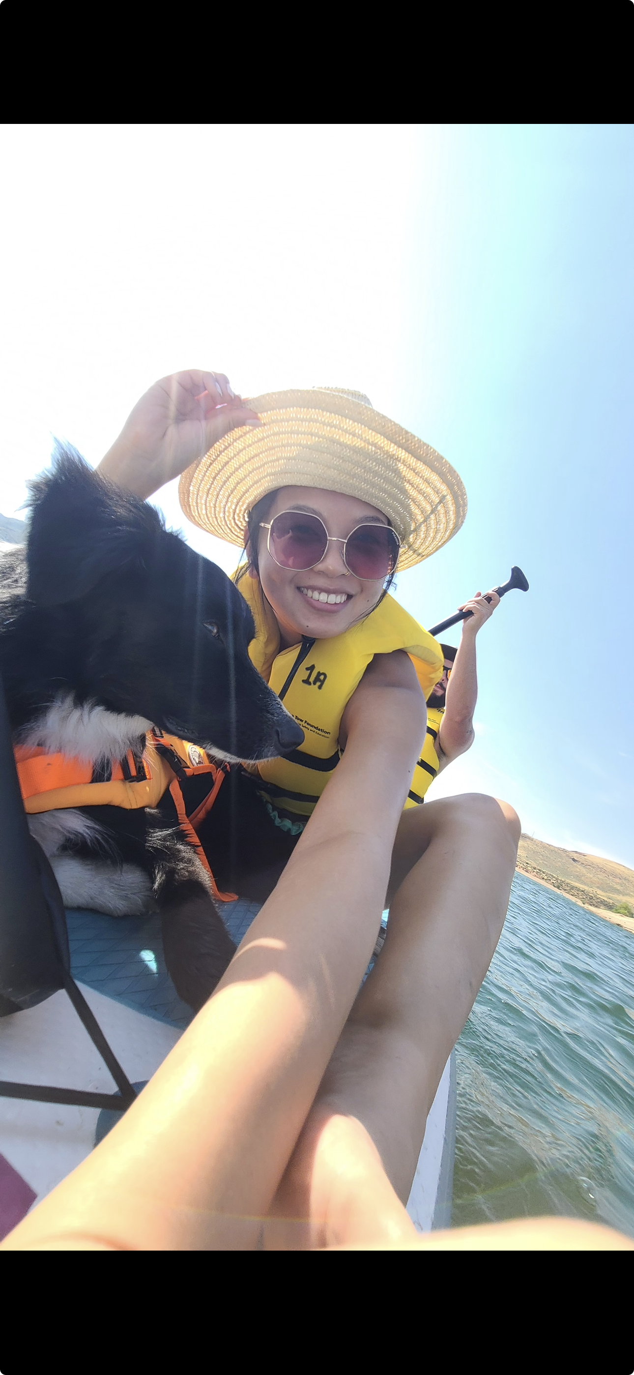 A woman with sunglasses and a straw hat smiling at the camera while sitting in a boat on a lake. She is wearing a yellow life jacket and holding a paddle, with a dog next to her. The background shows a clear sky and distant hills.